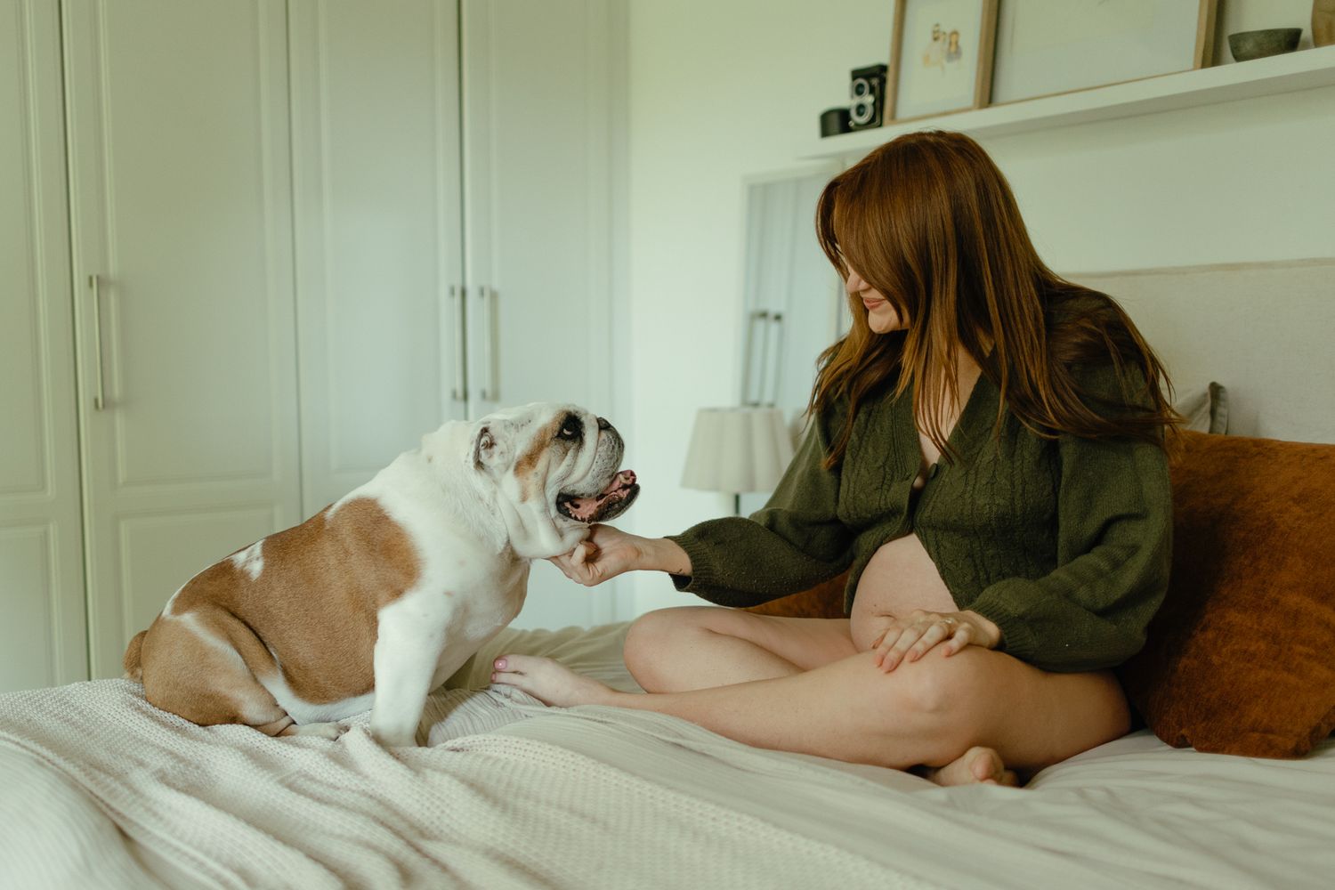 A pregnant woman sits cross-legged on a bed and reaches out to pet a friendly bulldog in a bright bedroom.