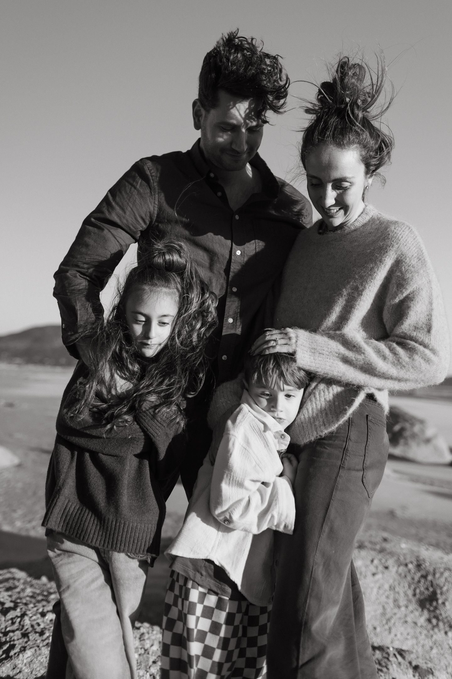Black and white photo of a laughing family of four hugging closely on a windswept beach.