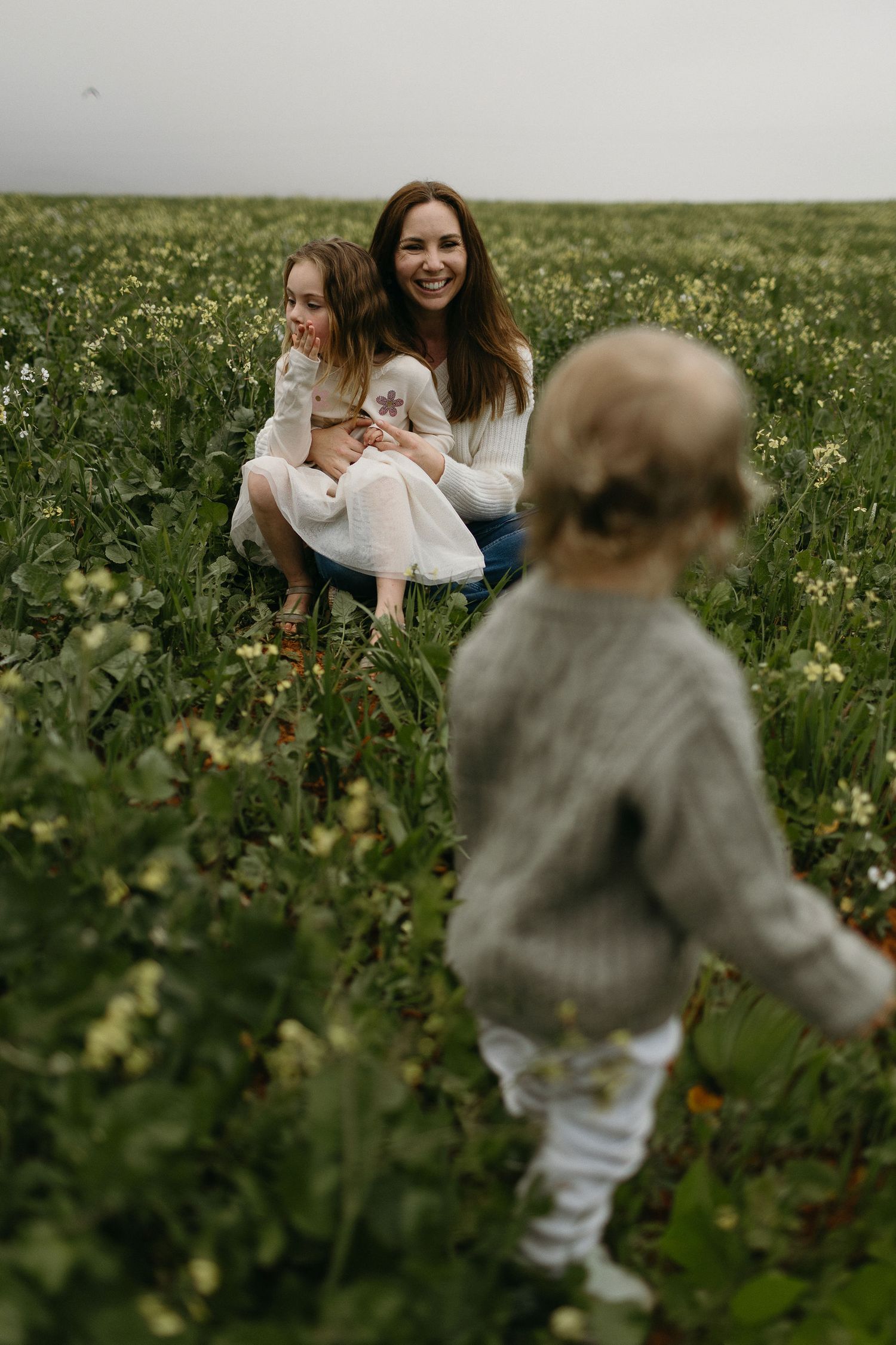 A smiling mother sits in a wildflower field with her daughter while a toddler in a knit sweater walks toward them.