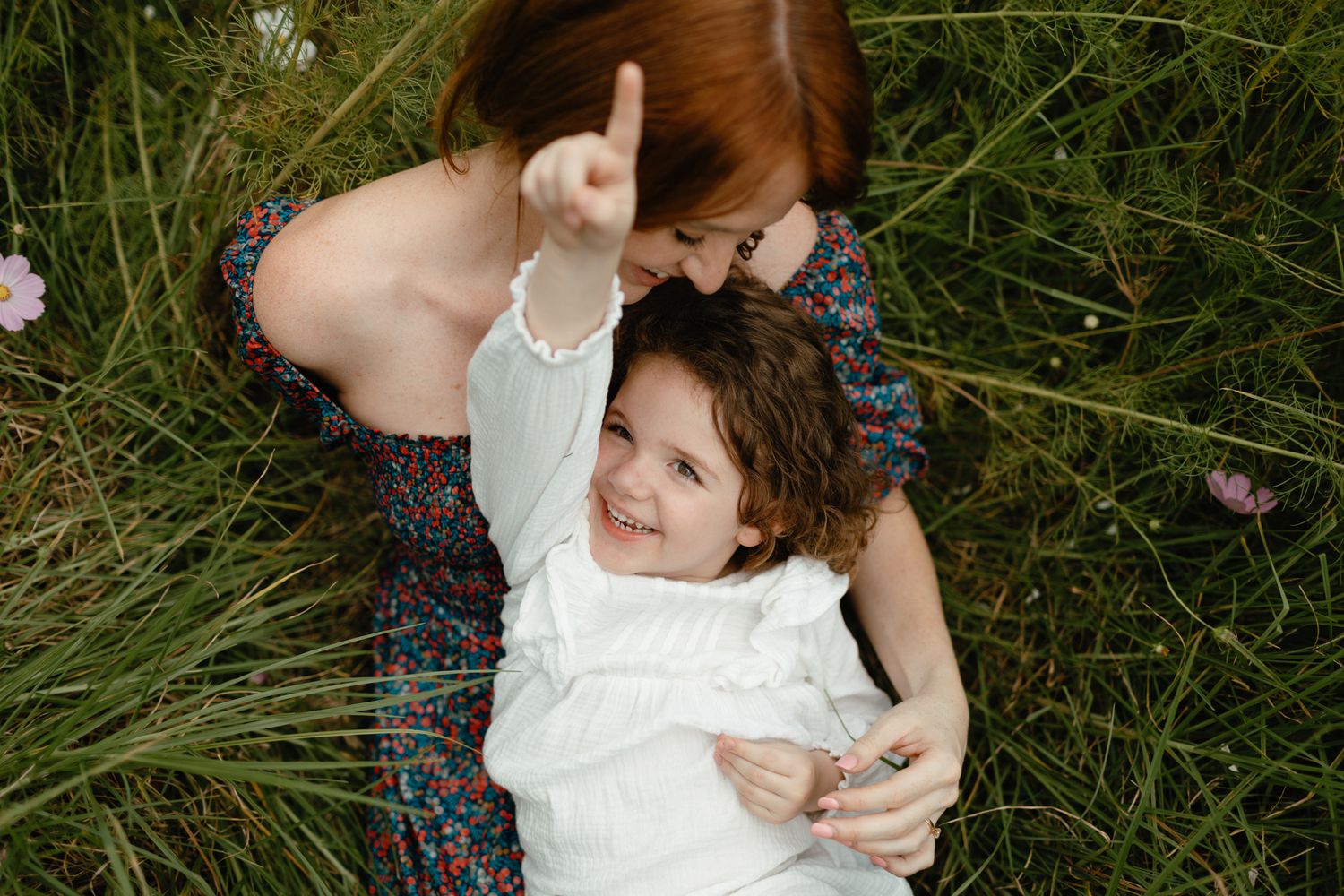 Joyful little girl points to the sky while lying in a wildflower meadow with her red-haired mother.