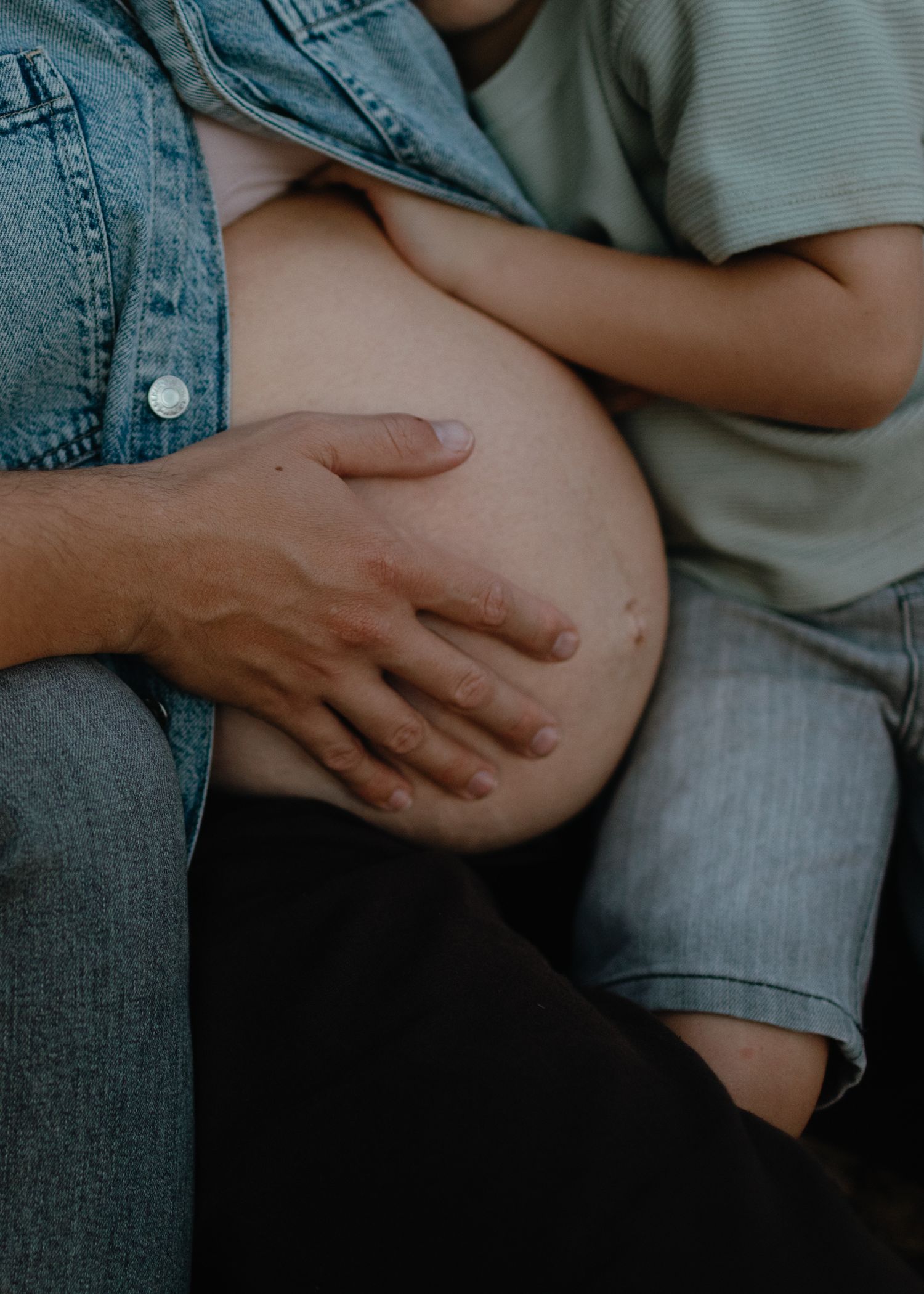 Close-up of a pregnant belly with a hand resting on it while a child in a green shirt leans in against the bump.