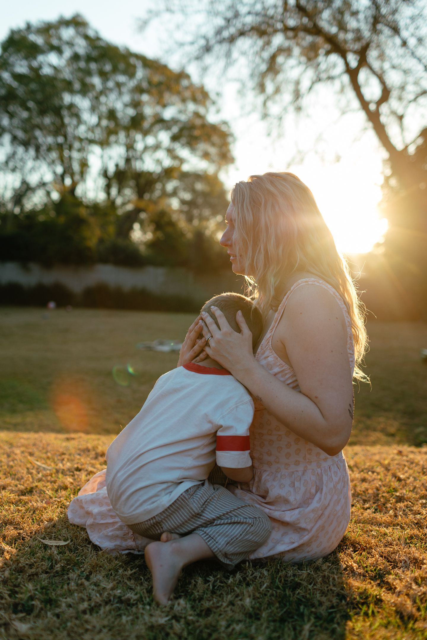 A blonde woman in a pink dress sits on dry grass cuddling a young child as golden sunset light glows through the trees.