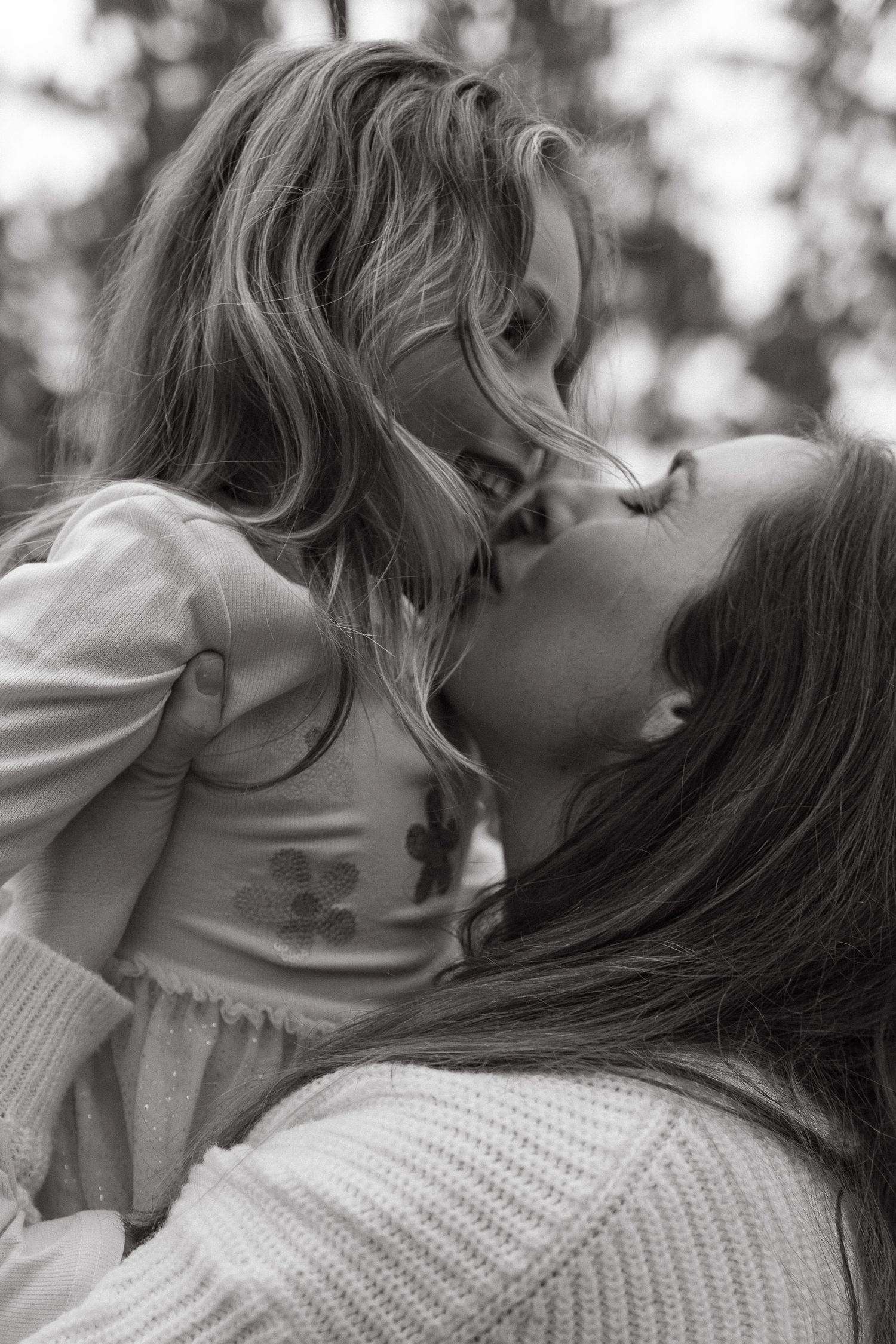 Black and white portrait of a mother kissing her smiling young daughter's cheek outdoors.