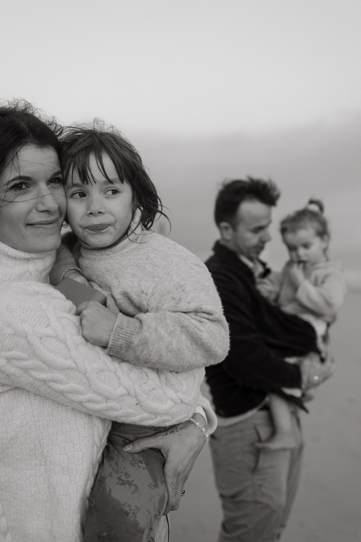 Black and white beach portrait of a mother holding her daughter while a father carries a toddler in soft fog.