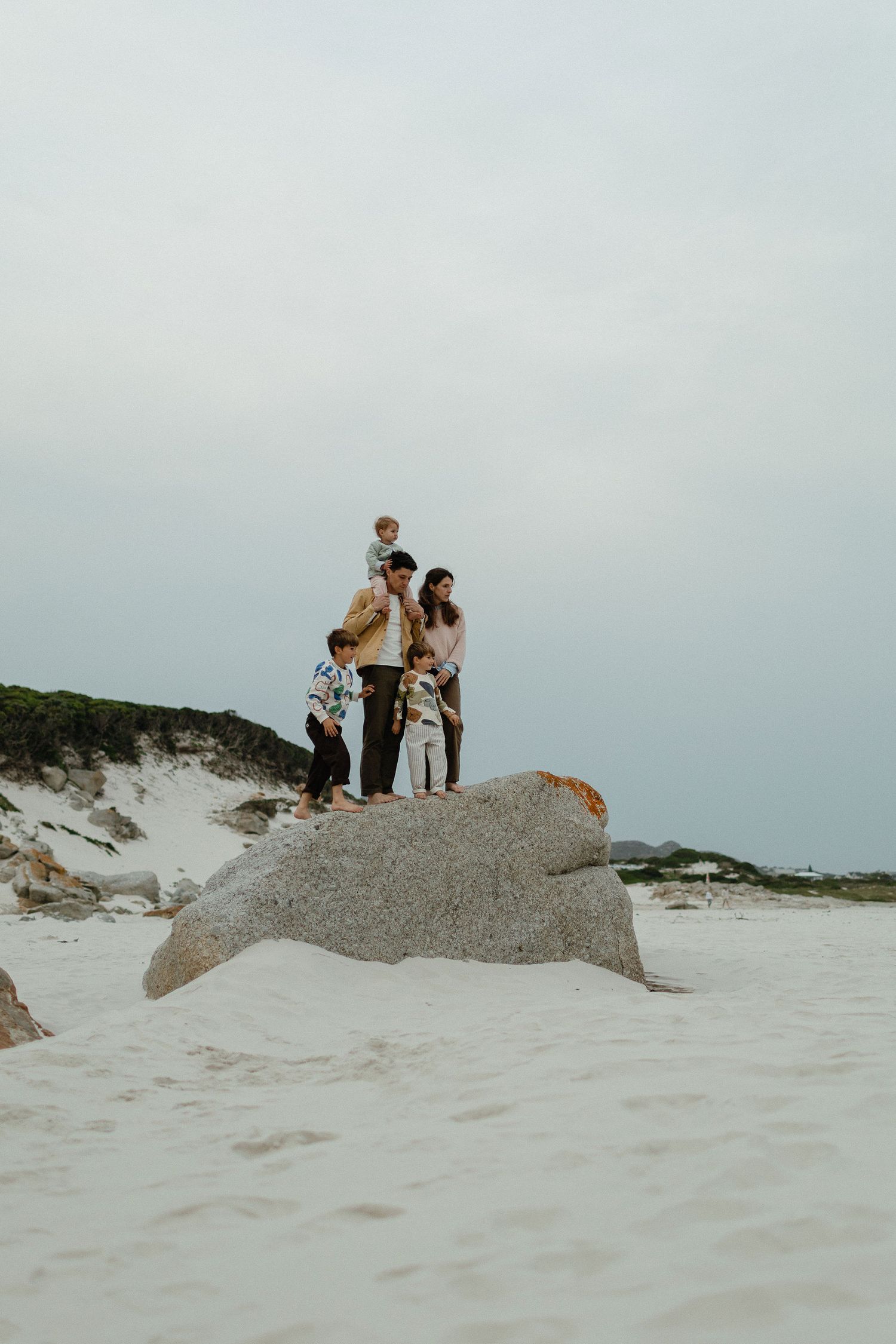 Family of five standing together on a large granite boulder on a white sand beach beneath a soft gray sky.