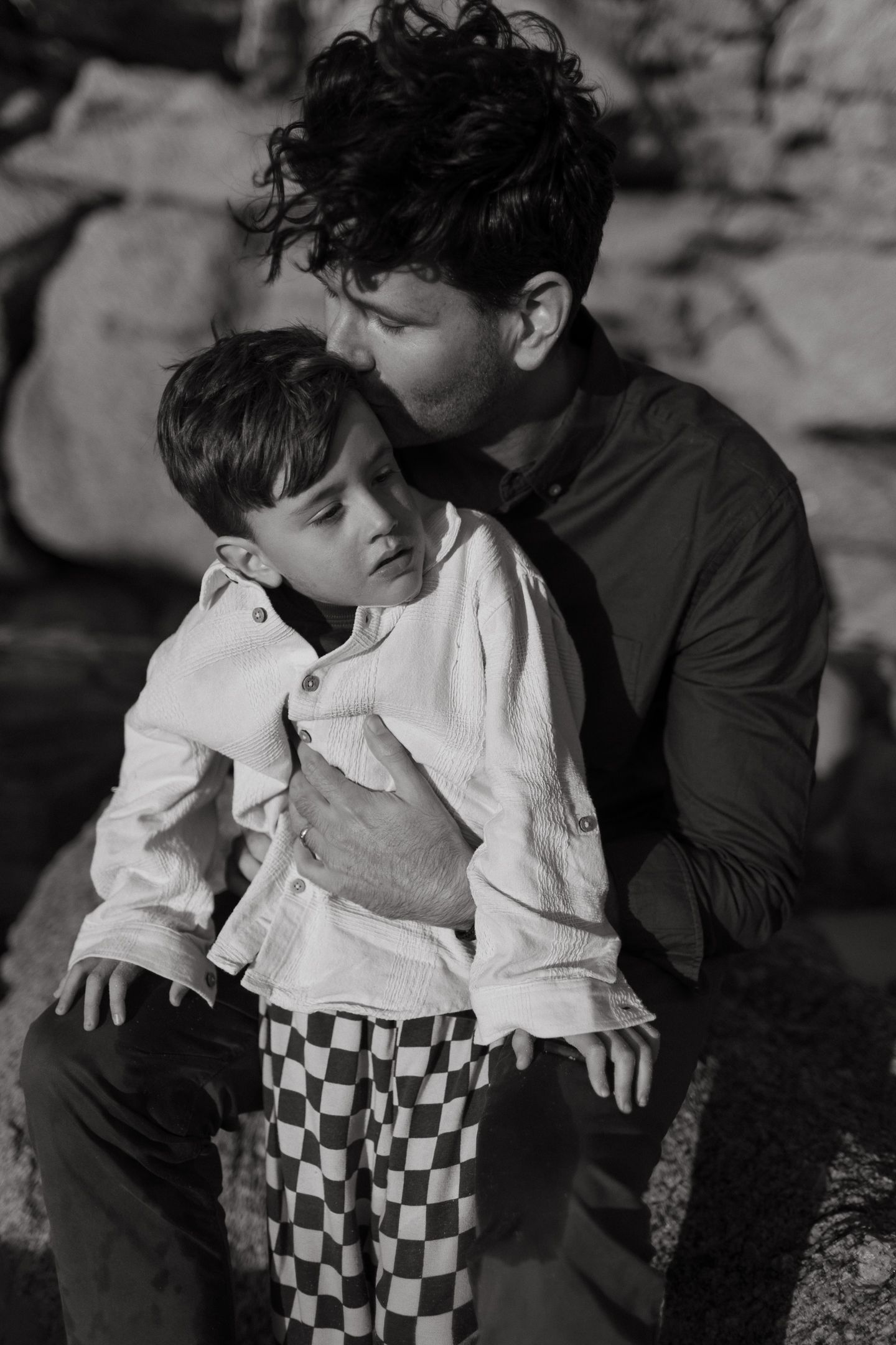 Black and white portrait of a father kissing his son's head as the boy sits on his lap in checkered pants.