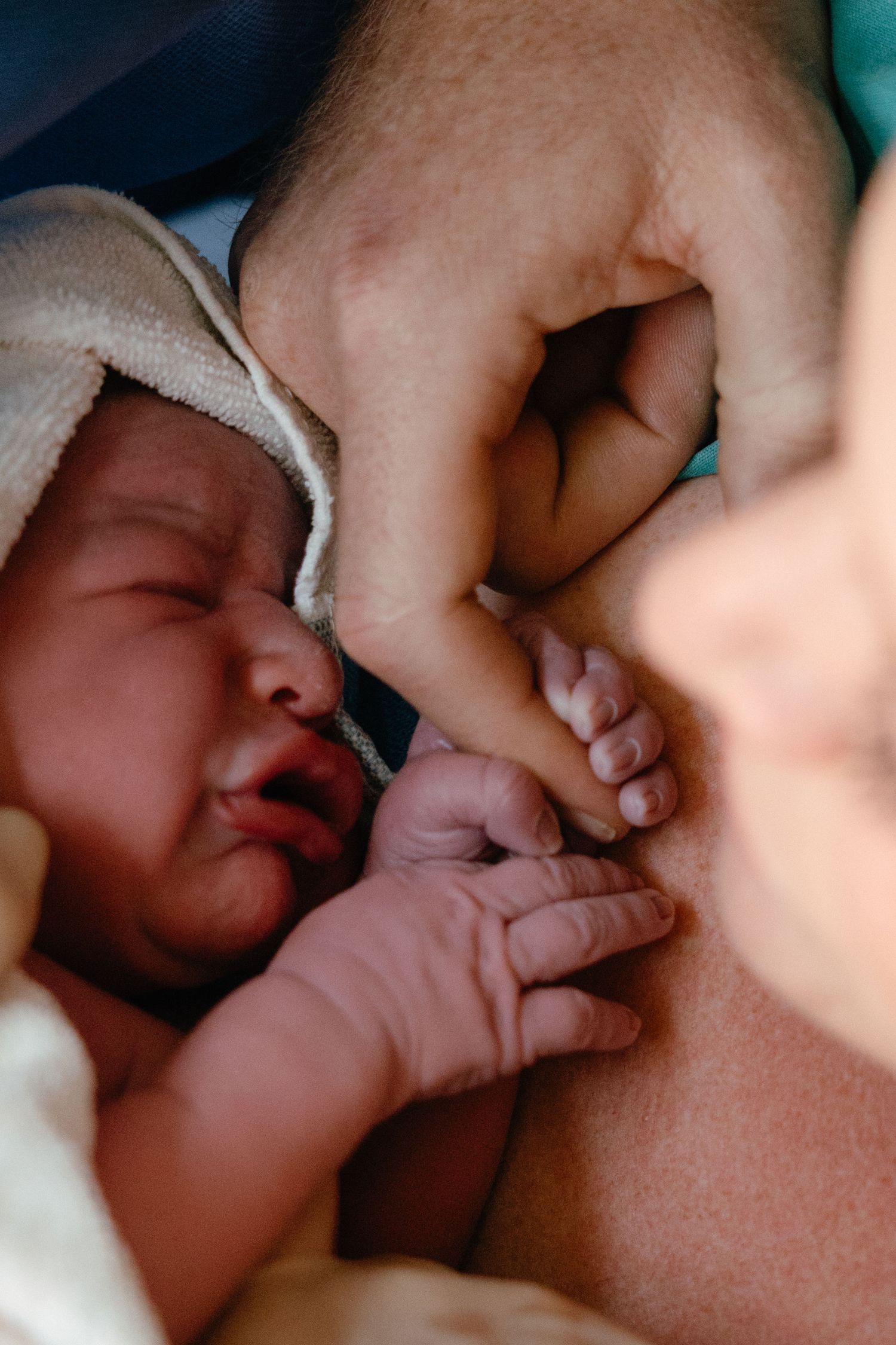 A crying newborn wrapped in a white blanket lies on the mother's chest while an adult hand gently cradles the baby's tiny fingers.