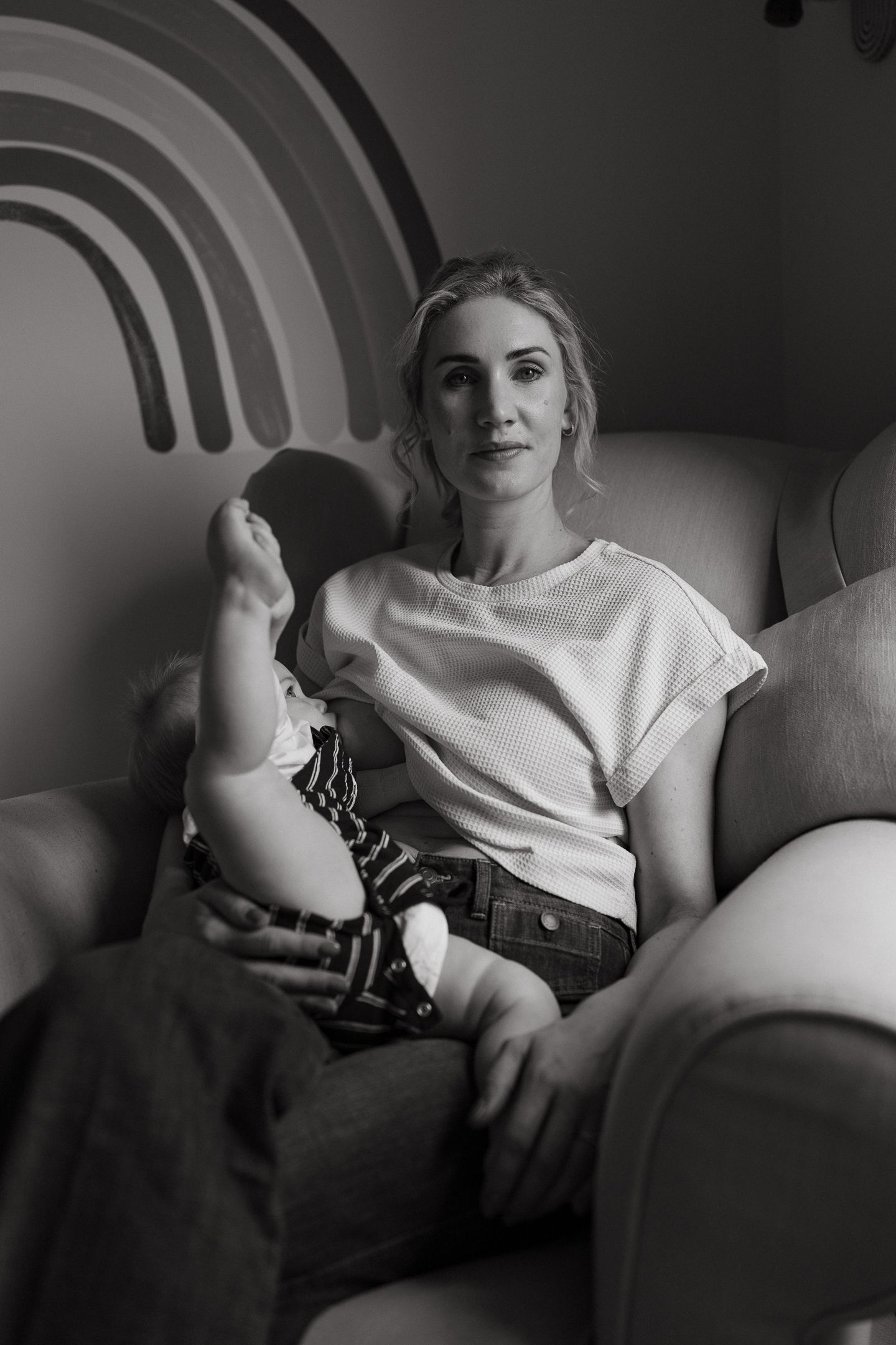 Black and white portrait of a mother seated in an armchair holding her baby in front of a rainbow wall mural.