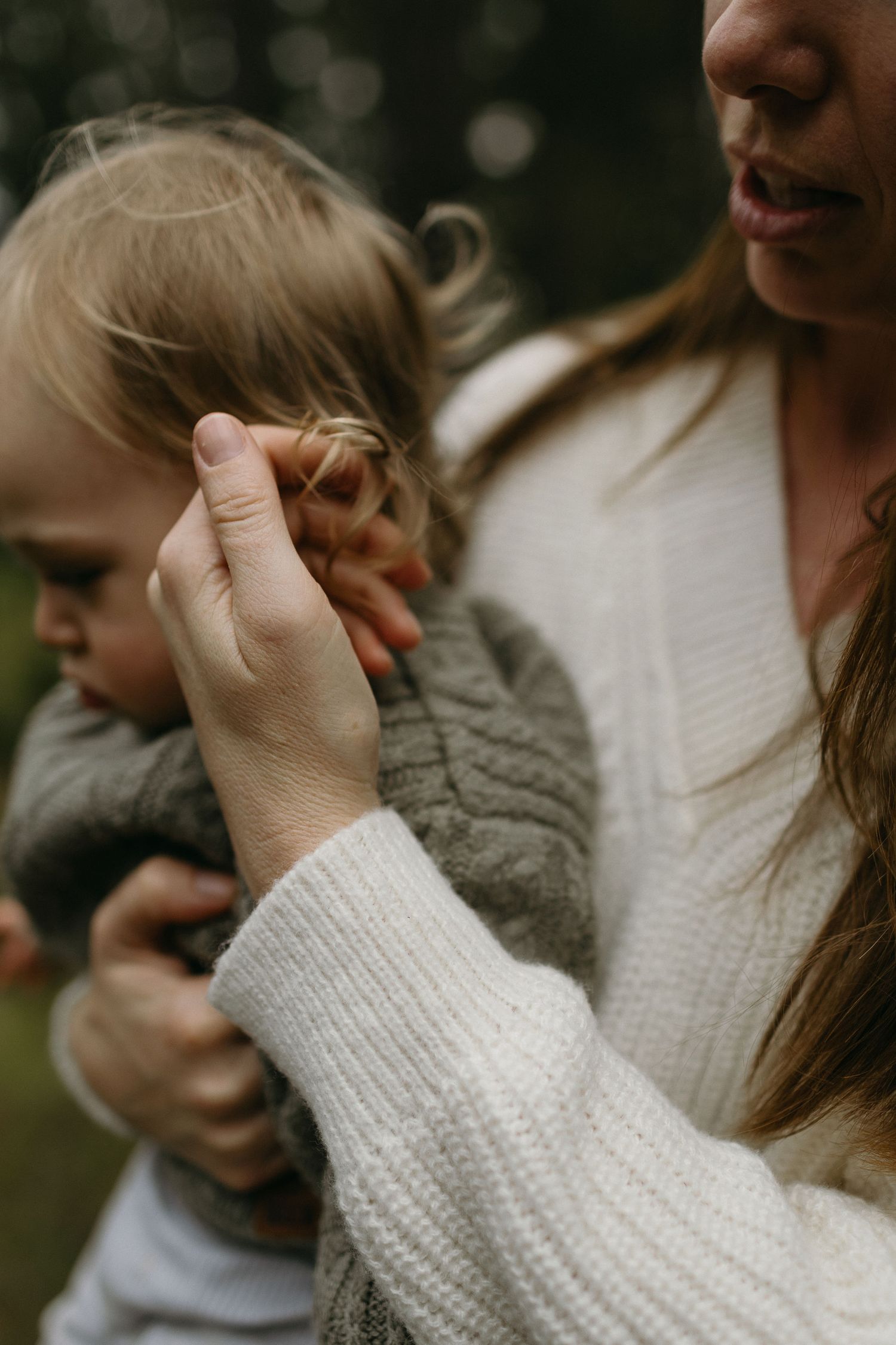 Mother in a white knit sweater tenderly touches the hair of her baby wrapped in a gray knit cardigan outdoors.