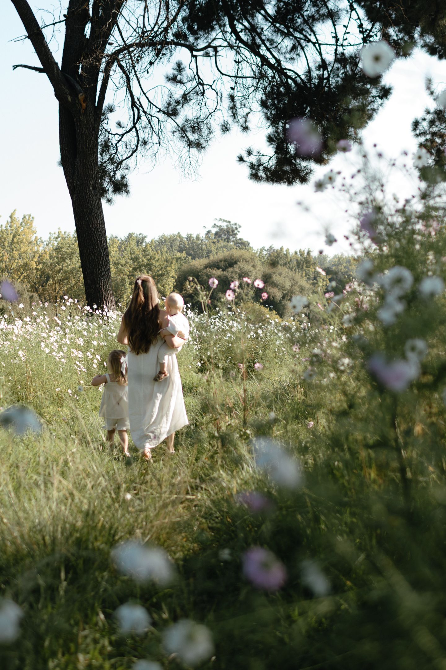 A mother in a white dress carries a toddler through a wildflower meadow while a young girl walks beside her under a tree.