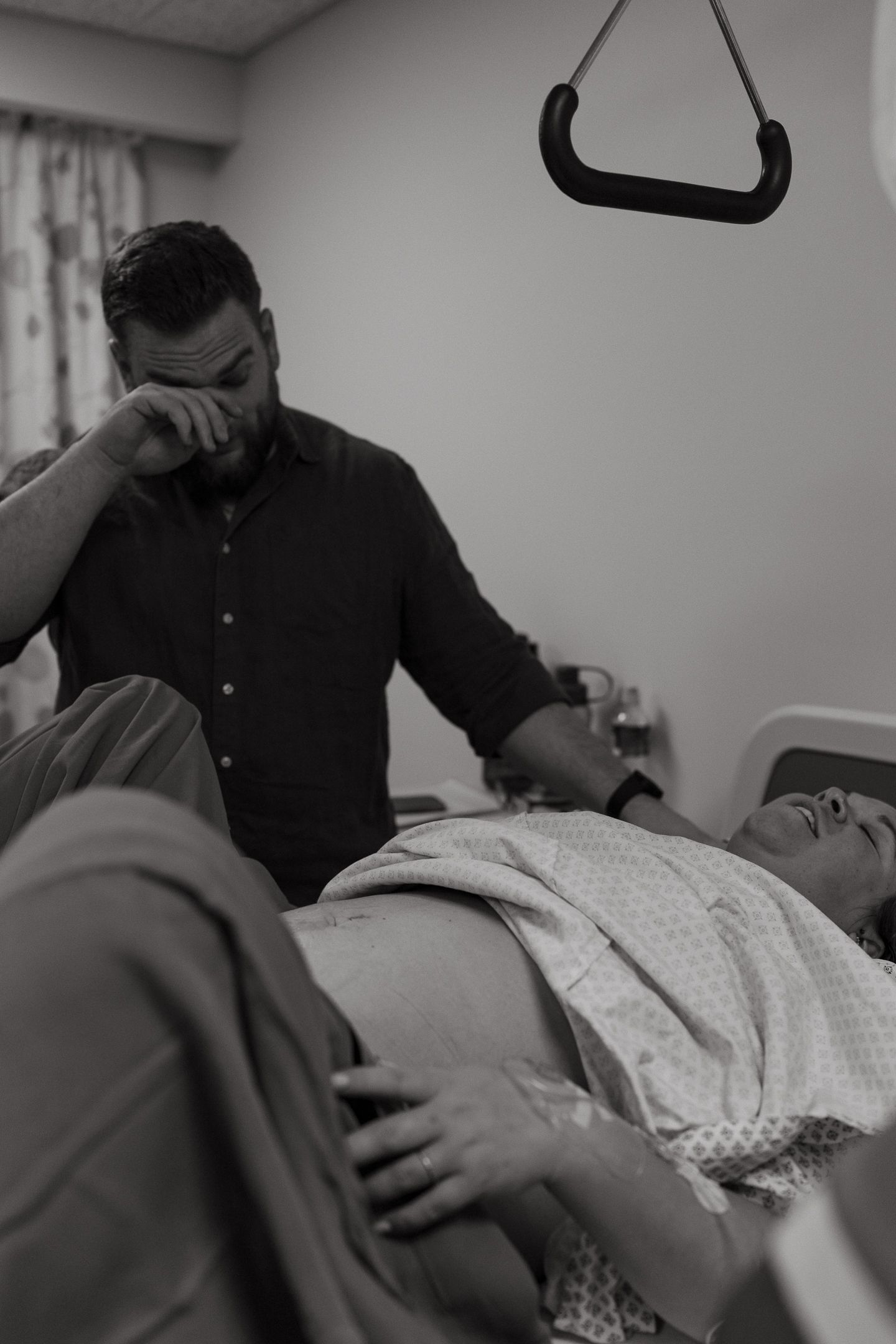 Emotional father wipes his eyes beside his wife lying in a hospital bed during labor and delivery.