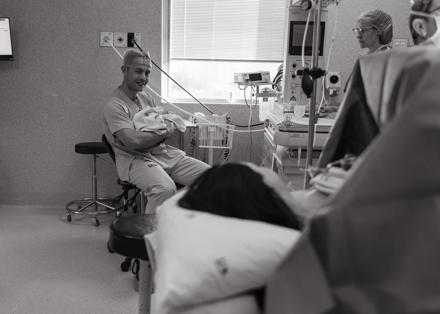 Smiling father in scrubs holds his newborn baby in a hospital room while the mother rests in bed and a nurse stands nearby.