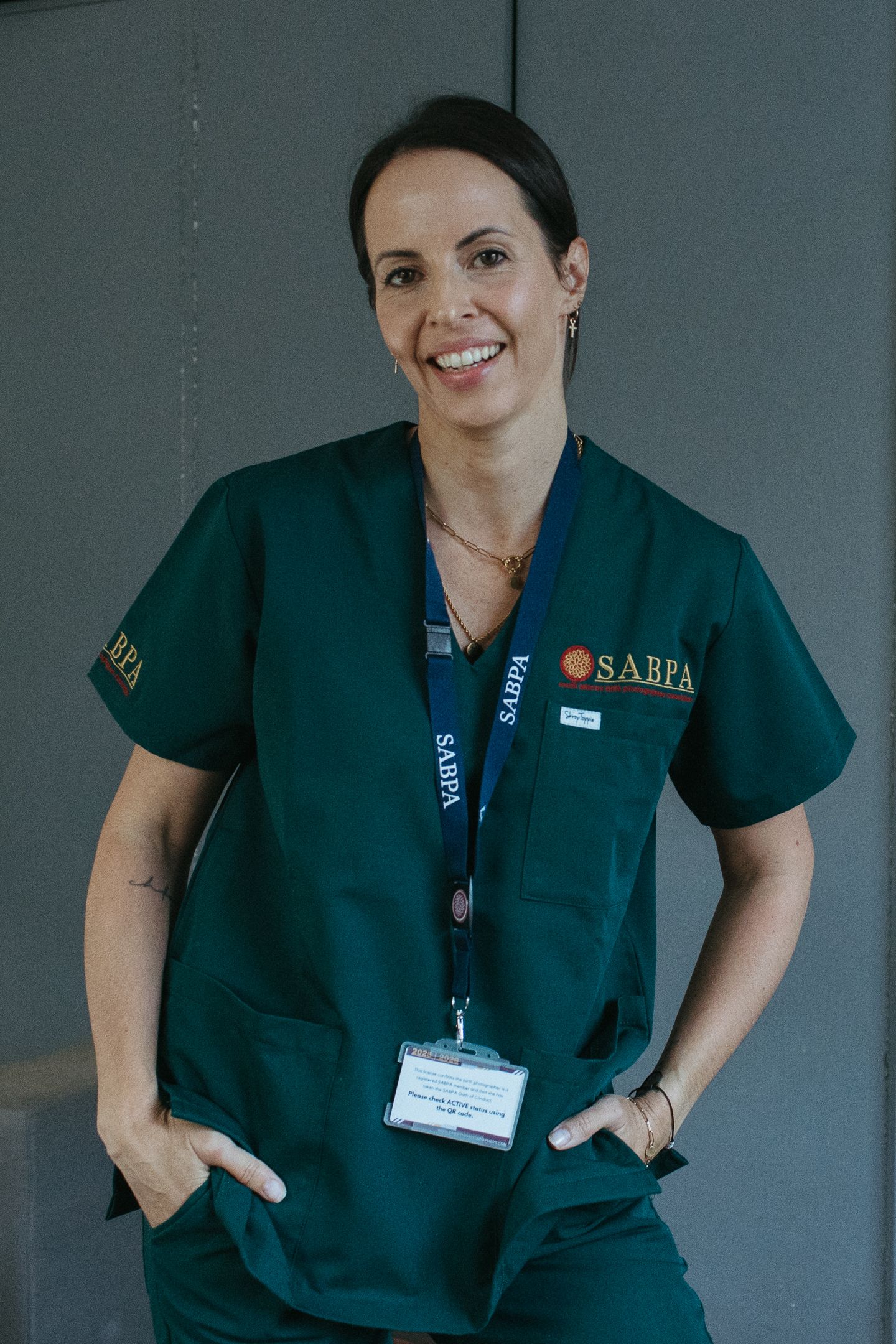 Smiling birth photographer in green SABPA scrubs and lanyard poses with hands in pockets against a gray wall in a hospital setting.