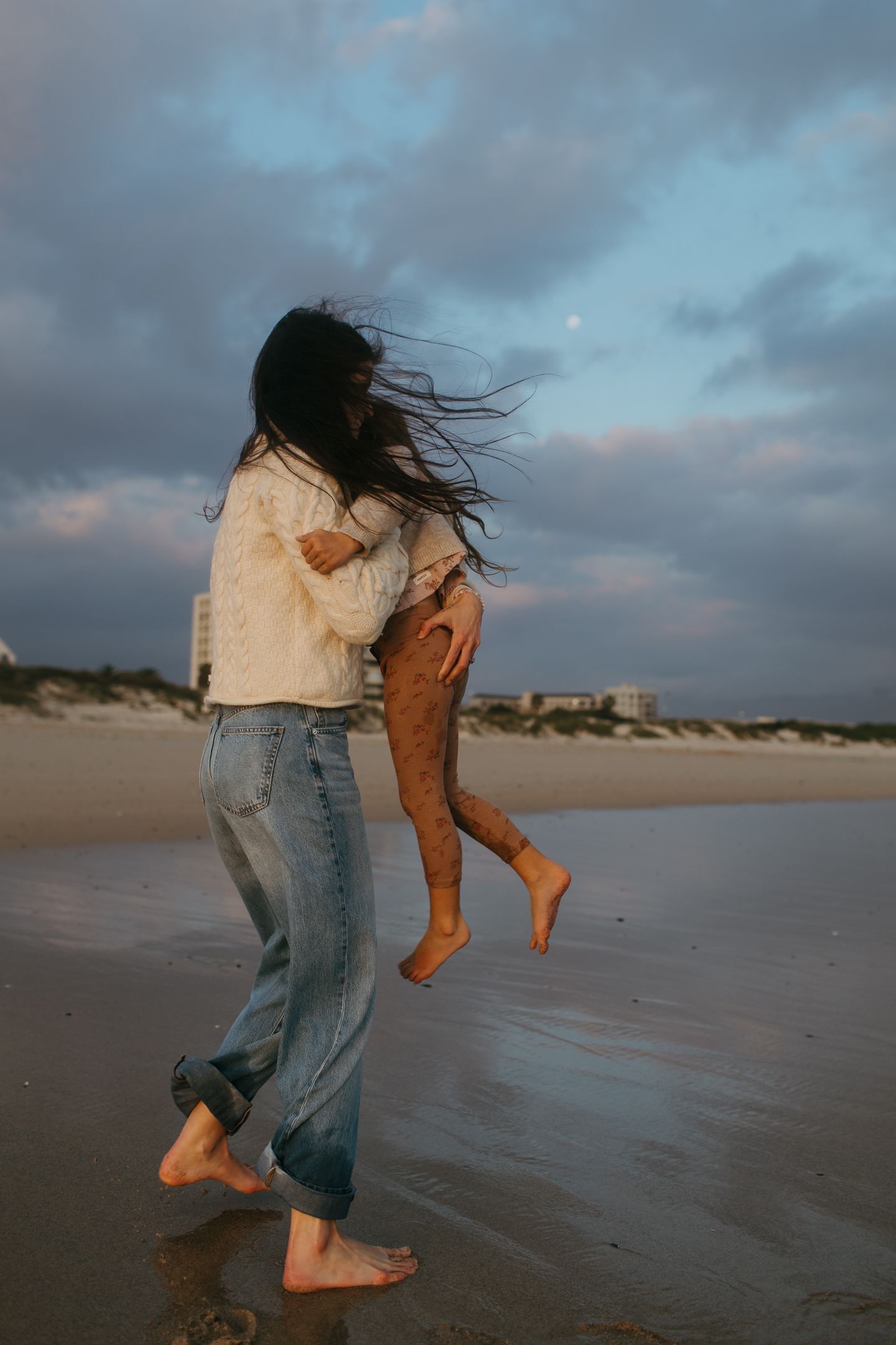 A barefoot woman in jeans lifts and carries a child along a wet beach under a cloudy sky as her hair blows in the wind.
