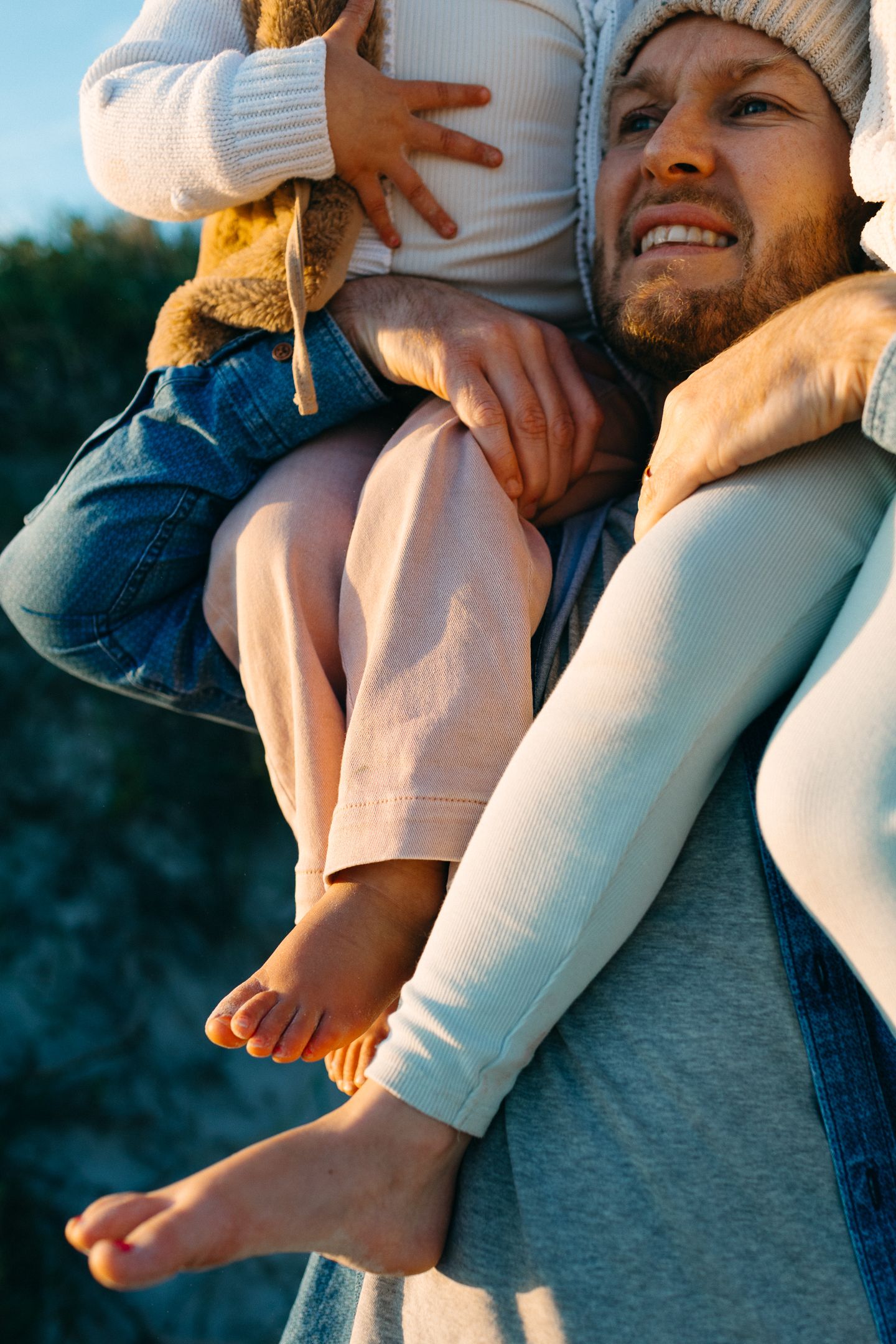 A bearded father in a knit beanie smiles while carrying a barefoot child on his shoulders outdoors in golden light.