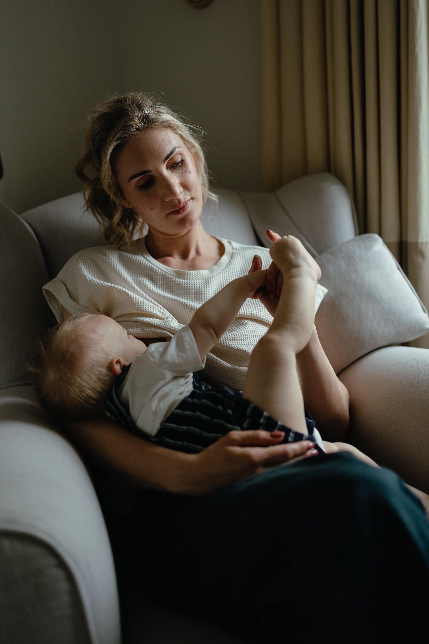 A mother smiles softly as she holds a baby who reaches up to play with her hand while she sits in a cozy armchair.