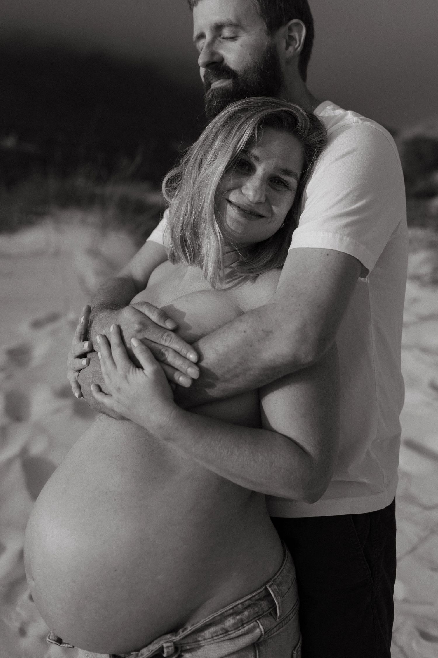 Black and white maternity photo of a bearded man embracing a pregnant woman from behind on a sandy beach.