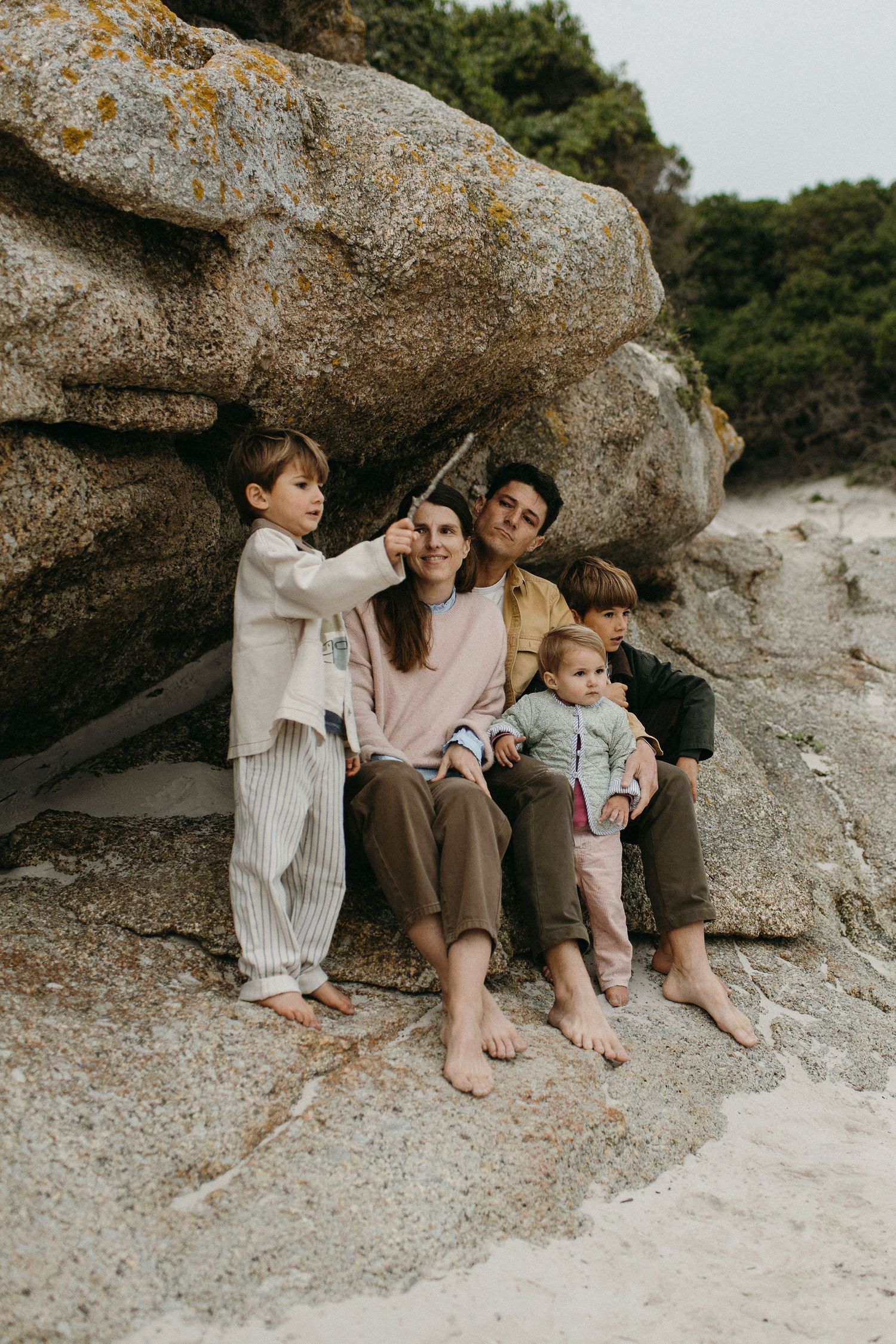 Smiling family with three young children sitting barefoot on large lichen-covered coastal boulders while a boy points toward the sky.