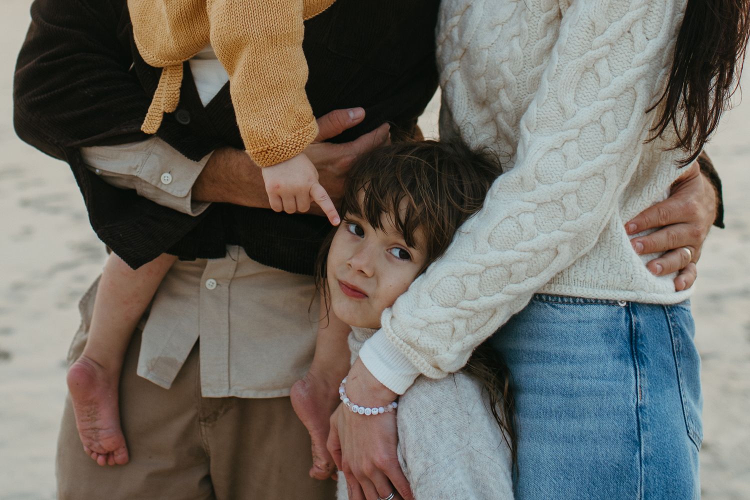 Young girl nestled between her parents on the beach while a toddler in a yellow cardigan is held in their arms.