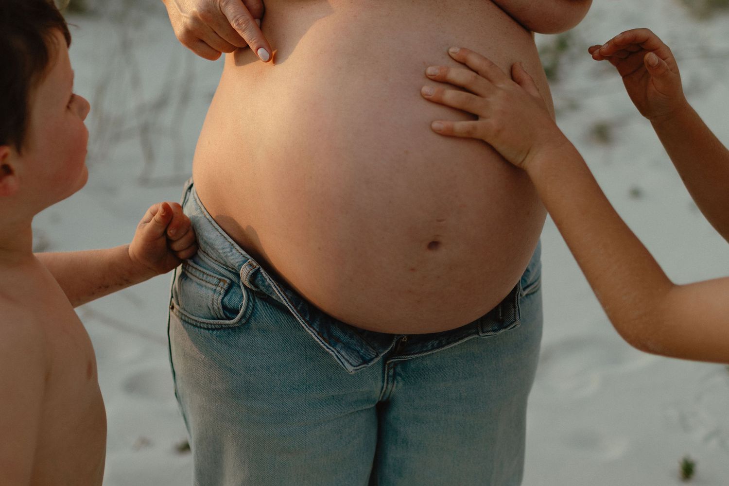 Two young children touch the bare pregnant belly of their mother wearing low-rise jeans on a sandy beach.