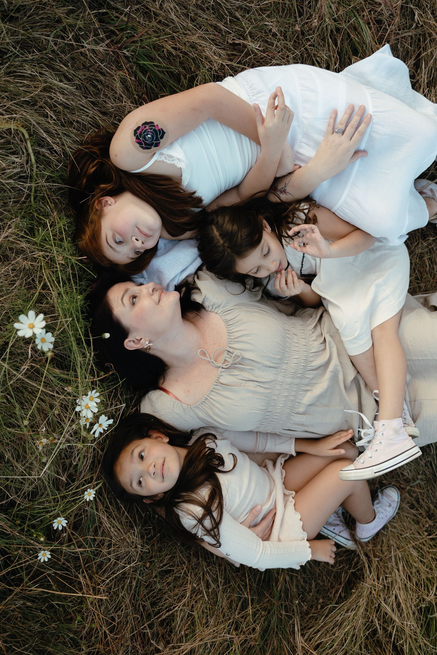 Overhead view of a mother and three daughters lying close together in dry grass beside small white daisies.