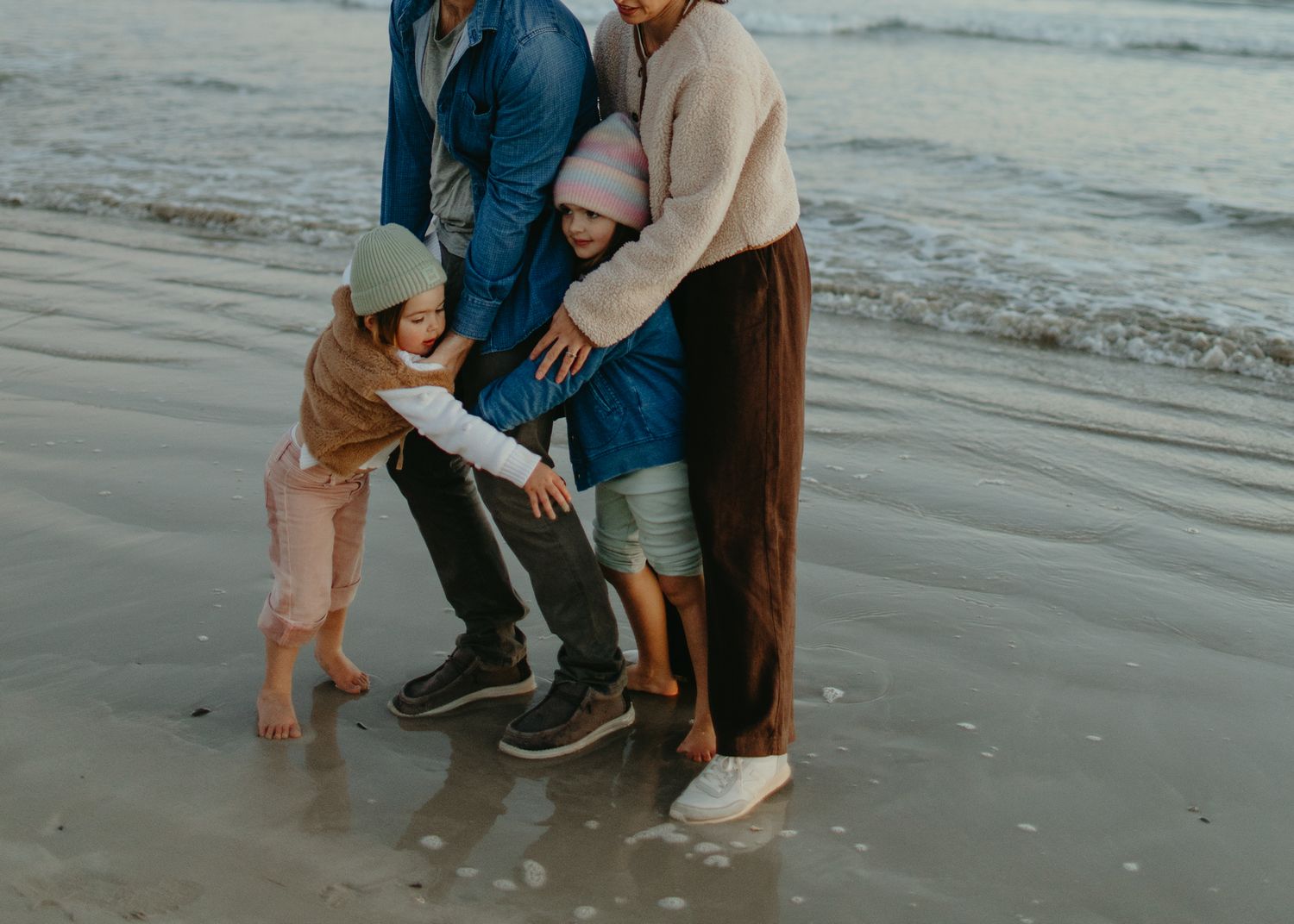 Family of four huddled together in a warm embrace on wet sand at the beach, with two children in beanies and ocean waves behind them.