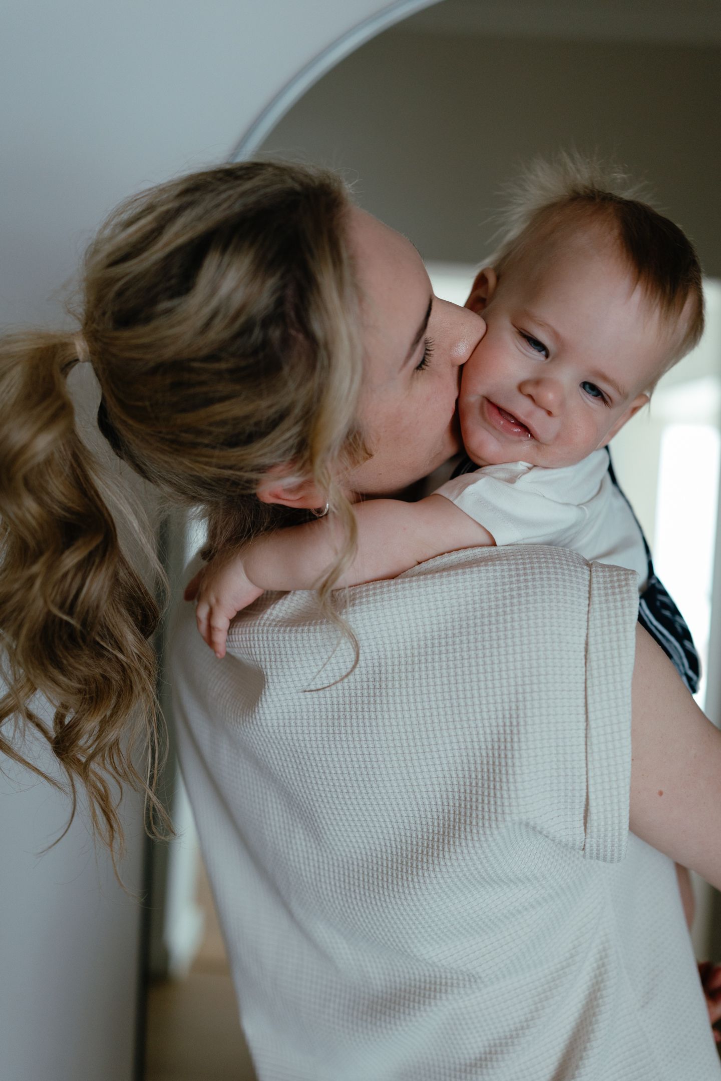 Close-up of a mother kissing her smiling baby on the cheek while holding him in a softly lit home.