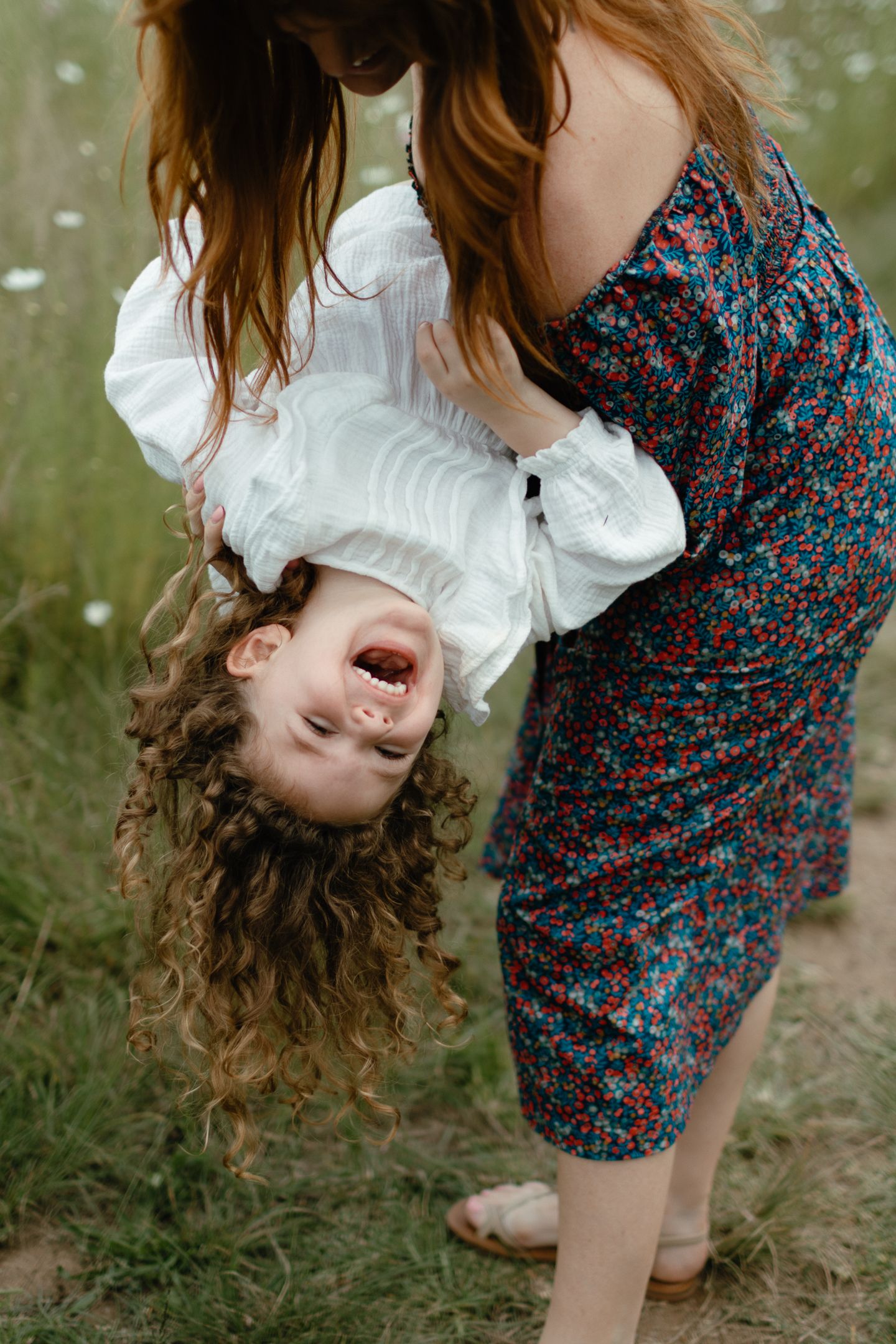 A smiling child with curly hair hangs upside down, laughing, held by a woman in a floral dress in a grassy field.