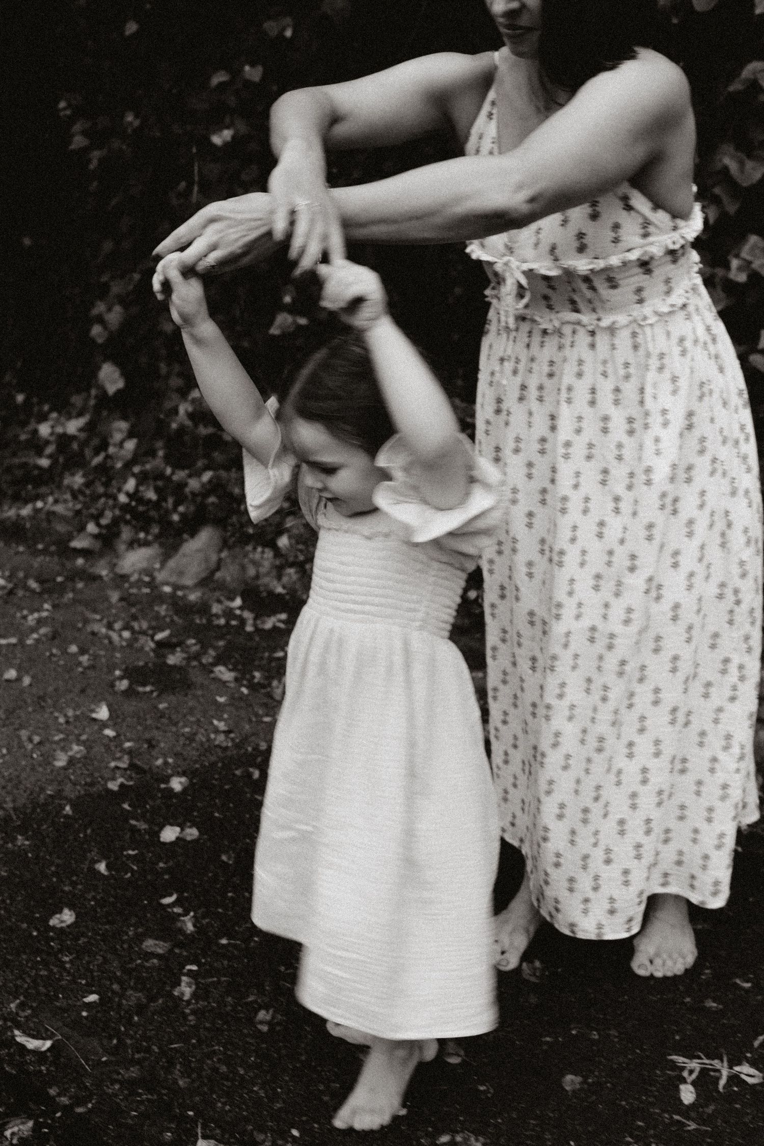 A black and white photo of a mother in a patterned dress holding hands with her young daughter as they dance outdoors.