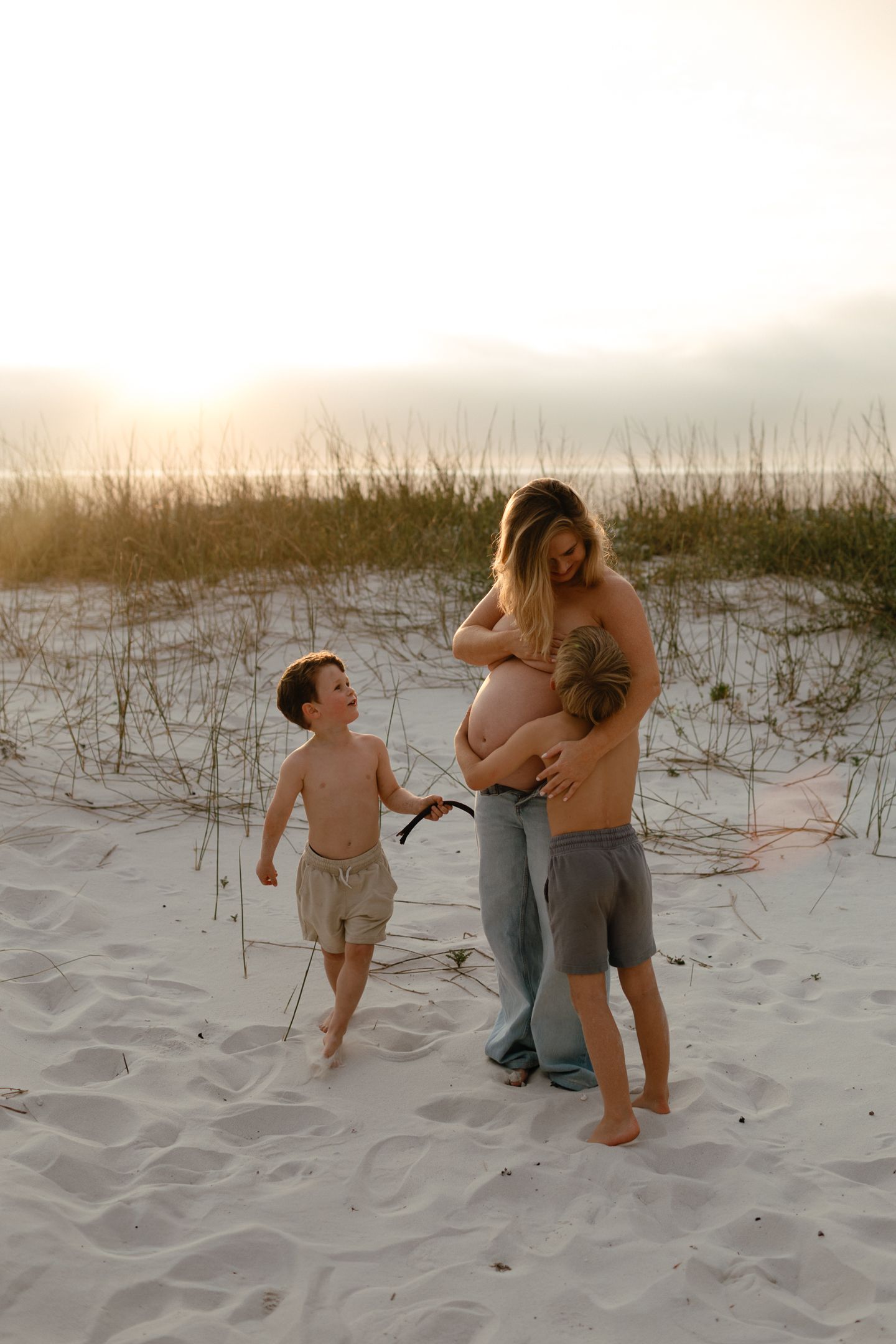 Pregnant mother and her two young sons share a tender moment on a sandy beach with dune grass at sunset.