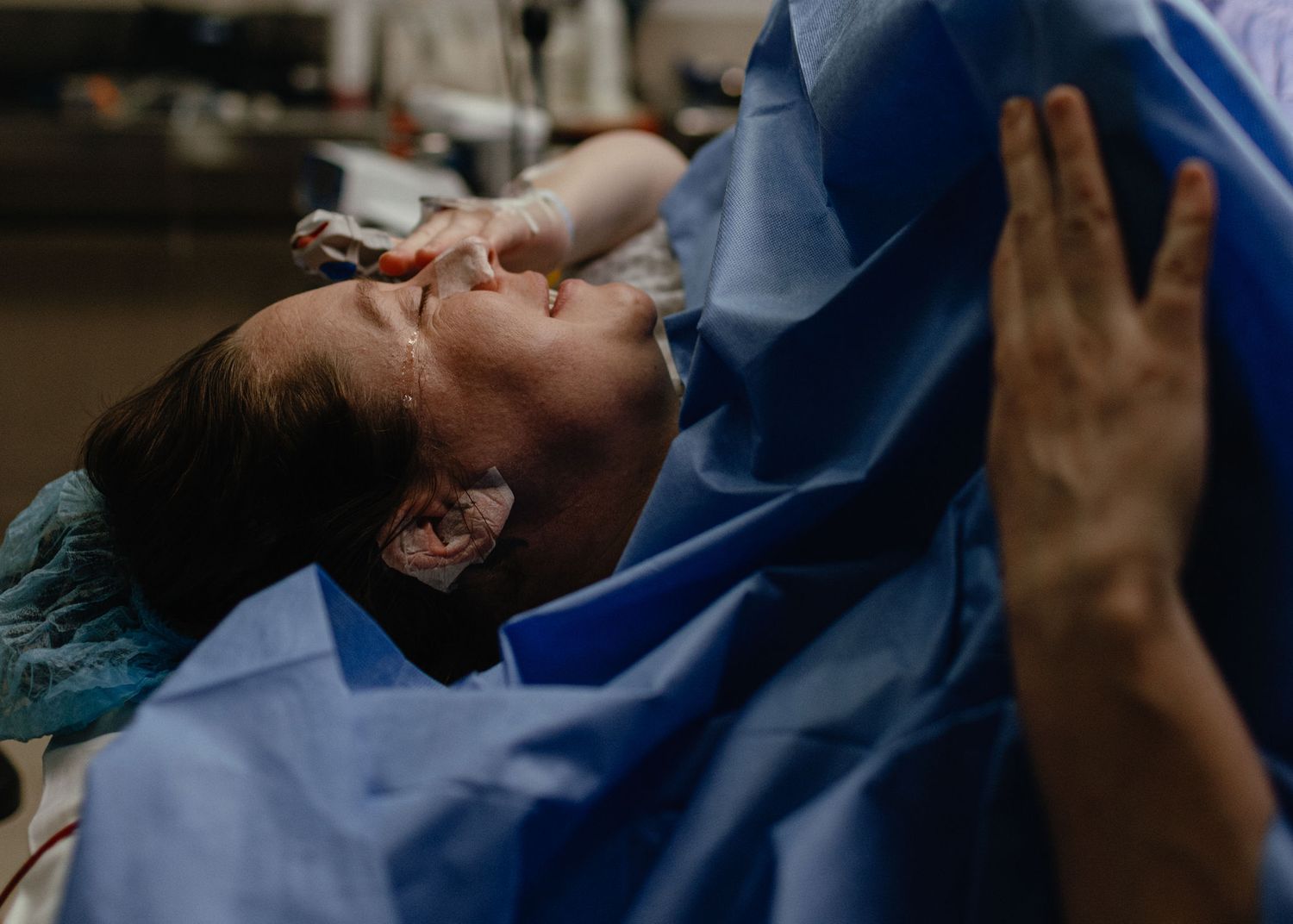 A woman in a blue hospital gown lies back with eyes closed and a calm expression during labor as a supportive hand rests beside her.