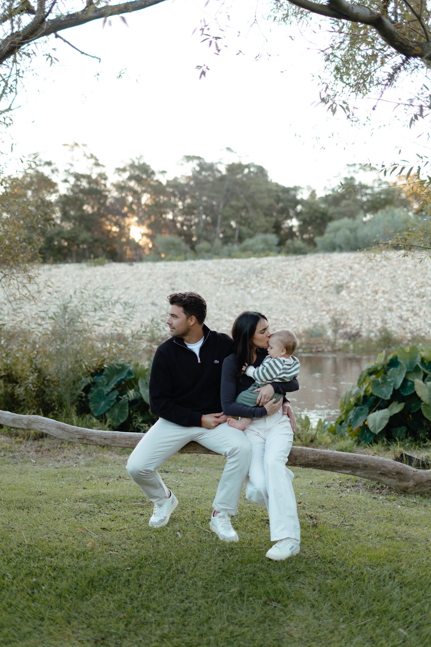 A couple sits on a fallen log with their baby on the grass beside a calm river at sunset.