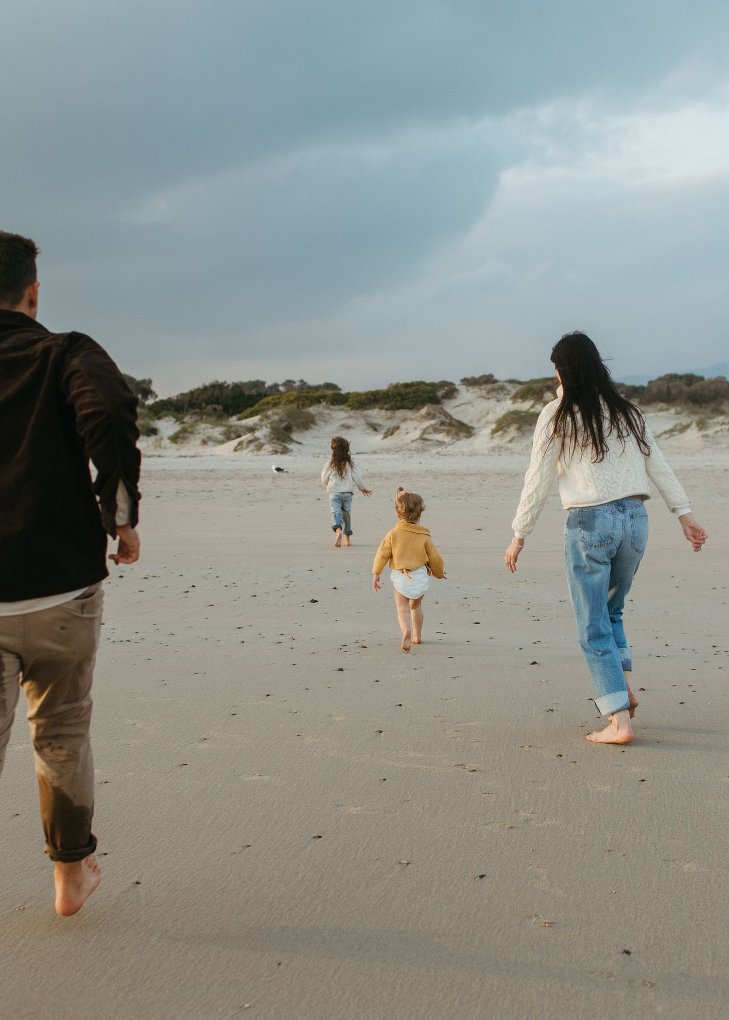 A family walks barefoot along a quiet beach toward dunes, with two children running ahead under moody skies.