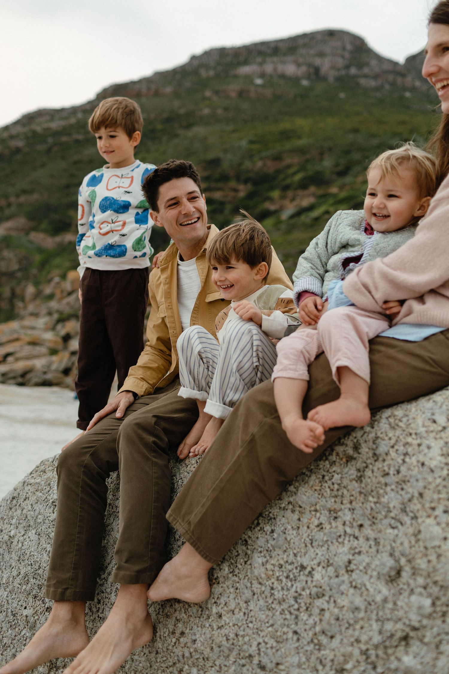 A smiling family with three young children sits barefoot on a large boulder near a grassy coastal hillside.