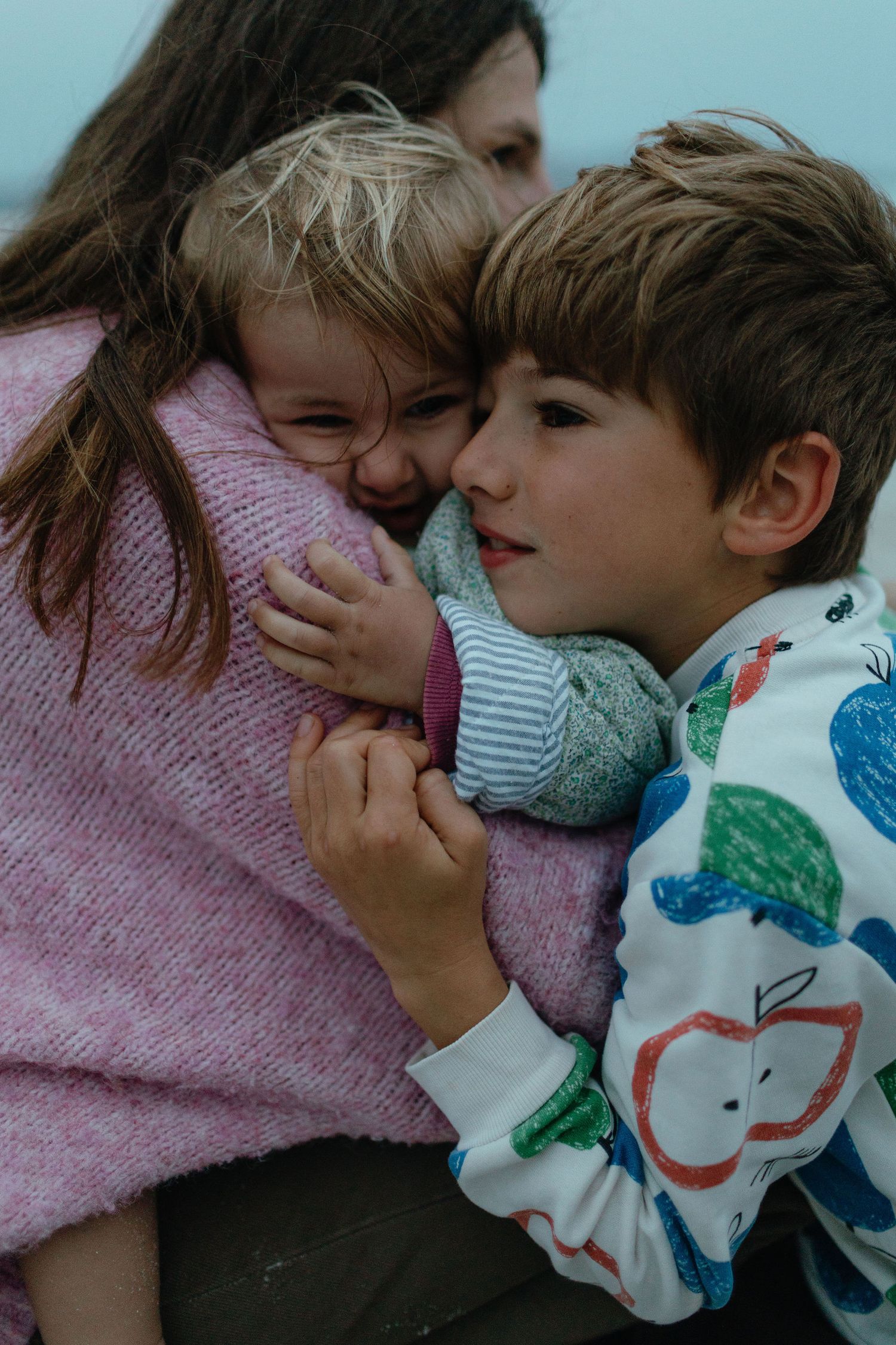 Two young brothers snuggle into their mother's pink sweater in a tender family hug on the beach.