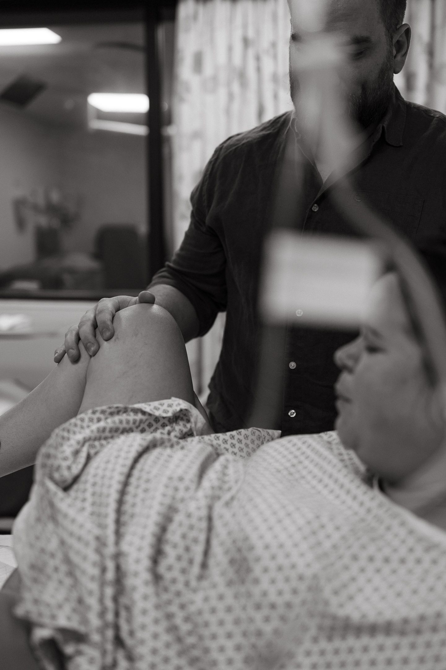 Black and white photo of a bearded man supporting a woman's raised leg as she lies in a hospital bed wearing a patterned gown during labor.