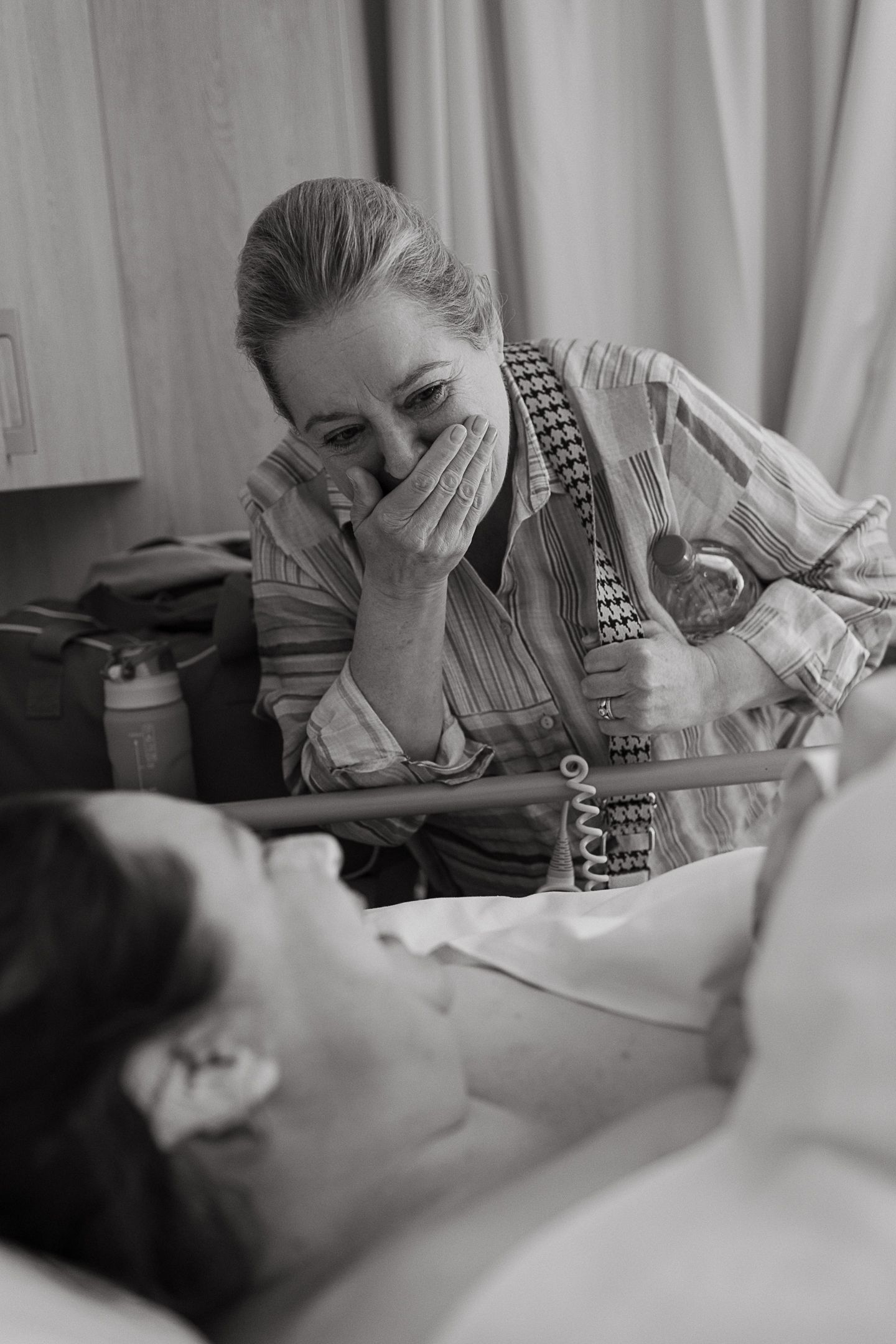Black and white photo of an emotional older woman covering her mouth with her hand as she looks at a patient lying in a hospital bed.