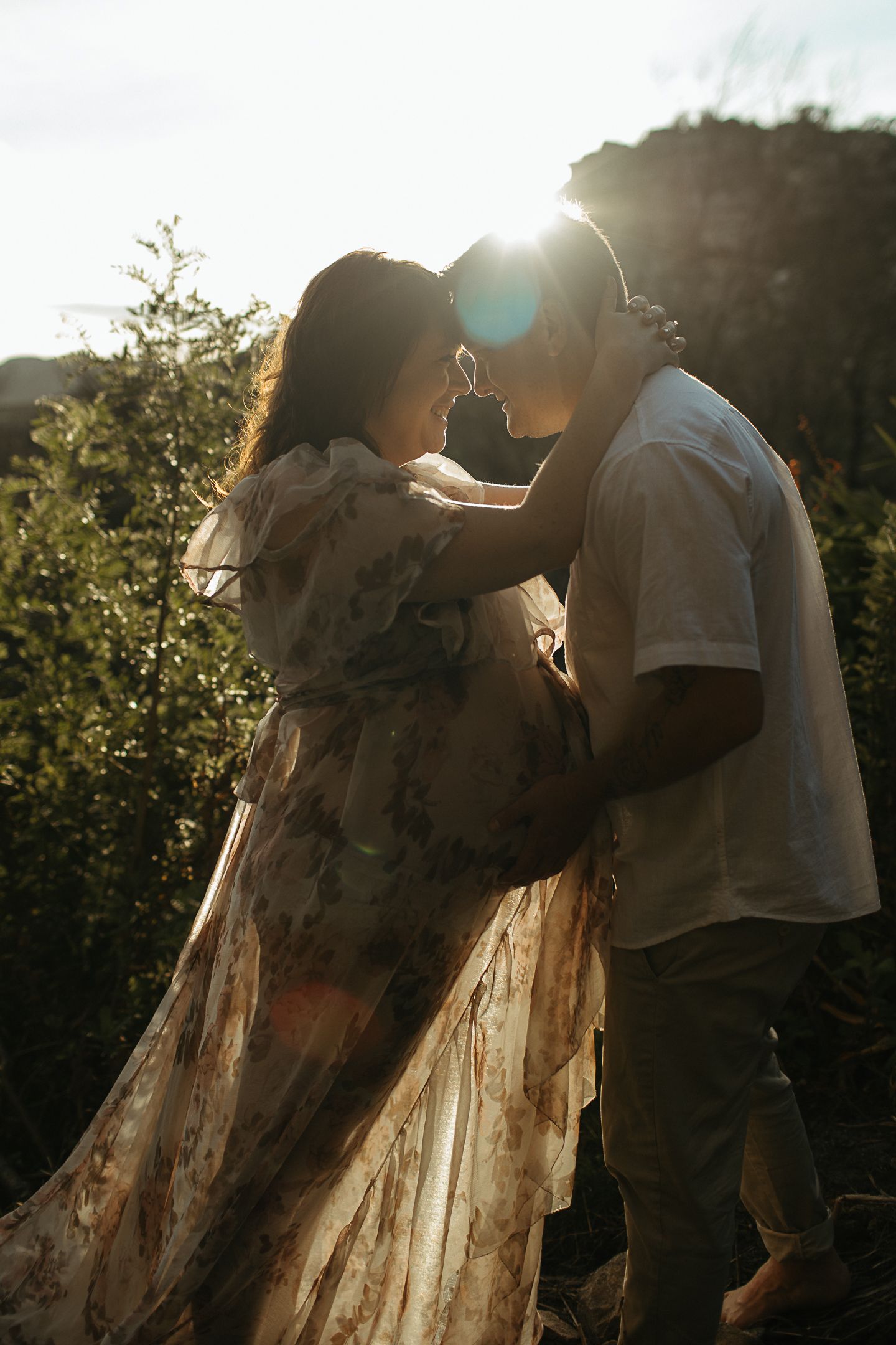 A silhouetted couple touches foreheads as the man cradles the pregnant belly of a woman in a floral gown at sunset.