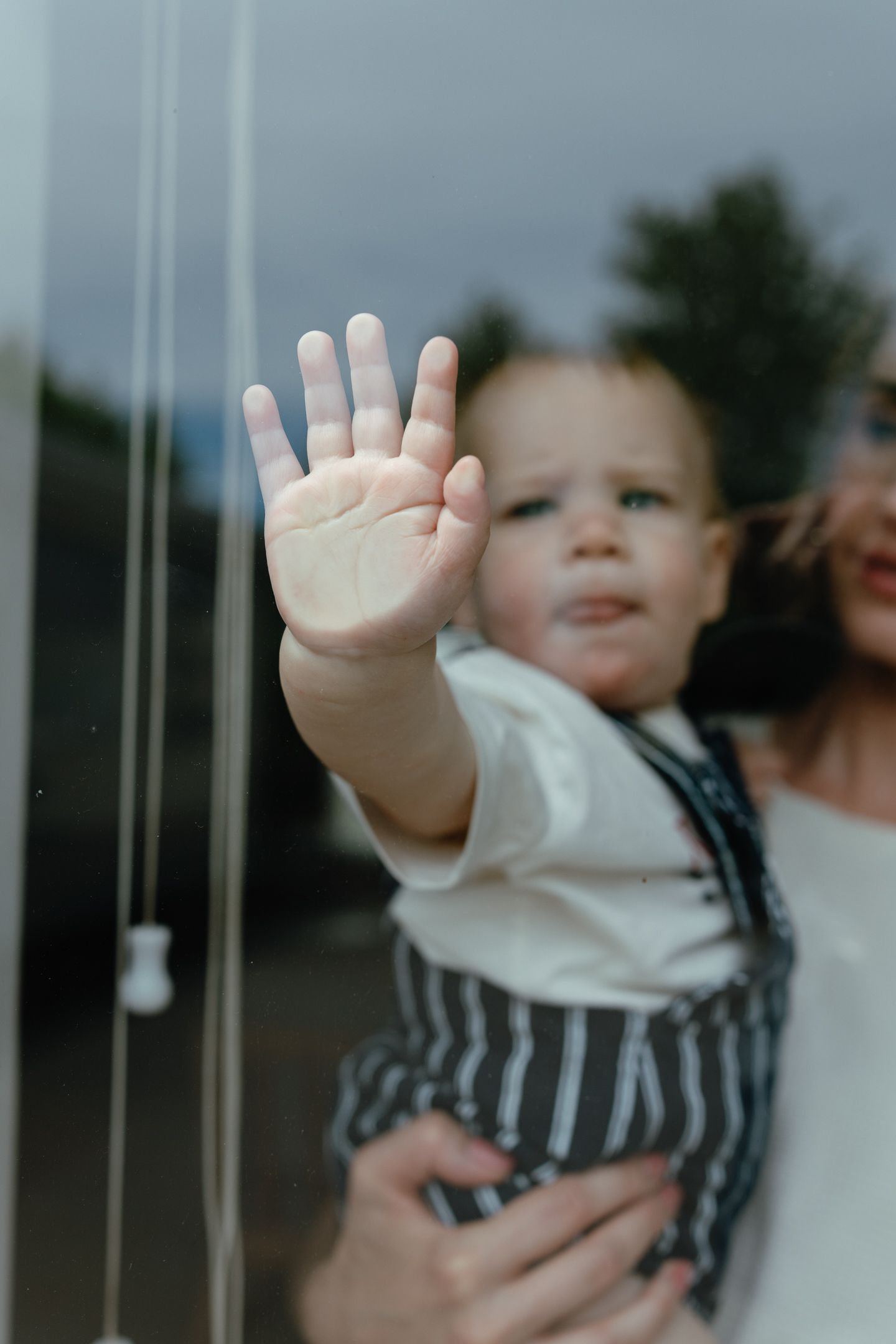 A baby's open hand presses against a window pane while his mother holds him, with trees reflected behind.