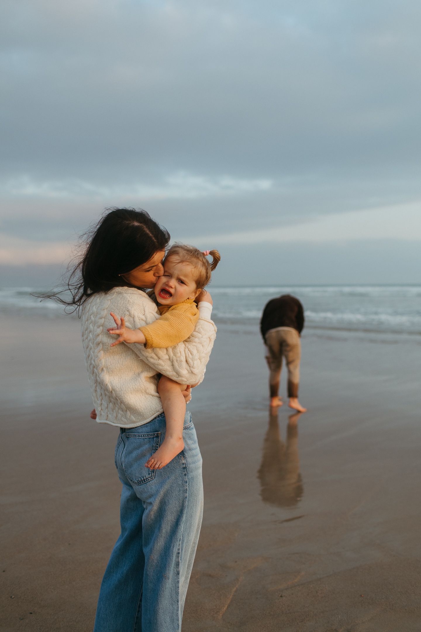 Mother in a cream sweater and jeans cuddling her toddler on a beach at dusk while another child plays near the waves.