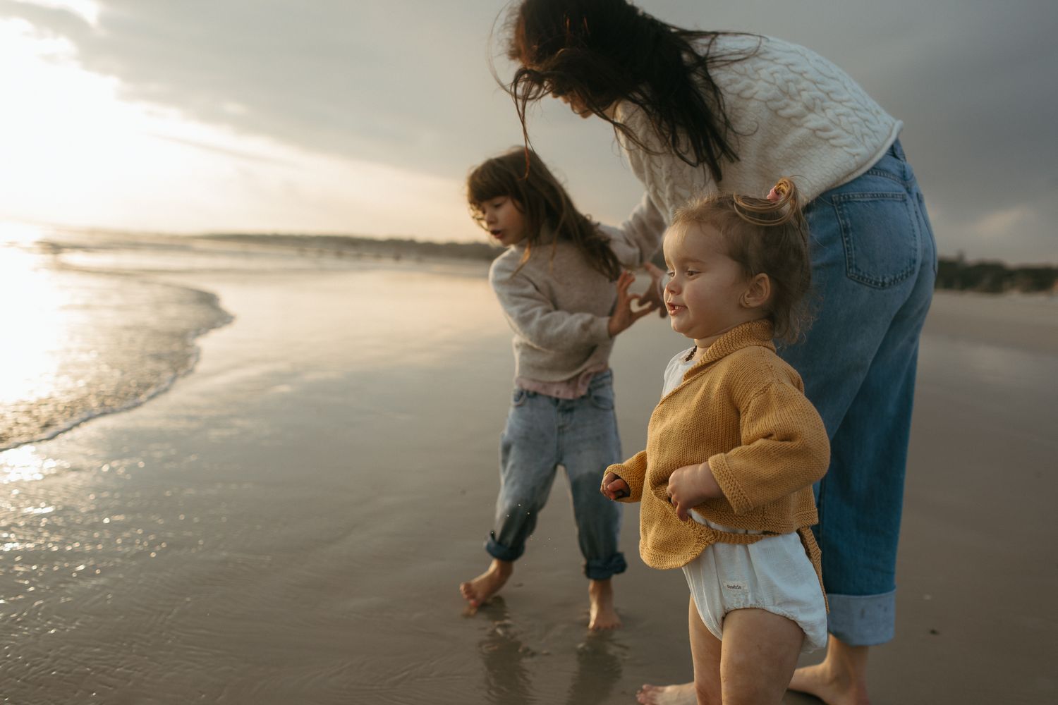 Mother crouches beside a young girl and a toddler in a yellow cardigan as they enjoy the shoreline at sunset.