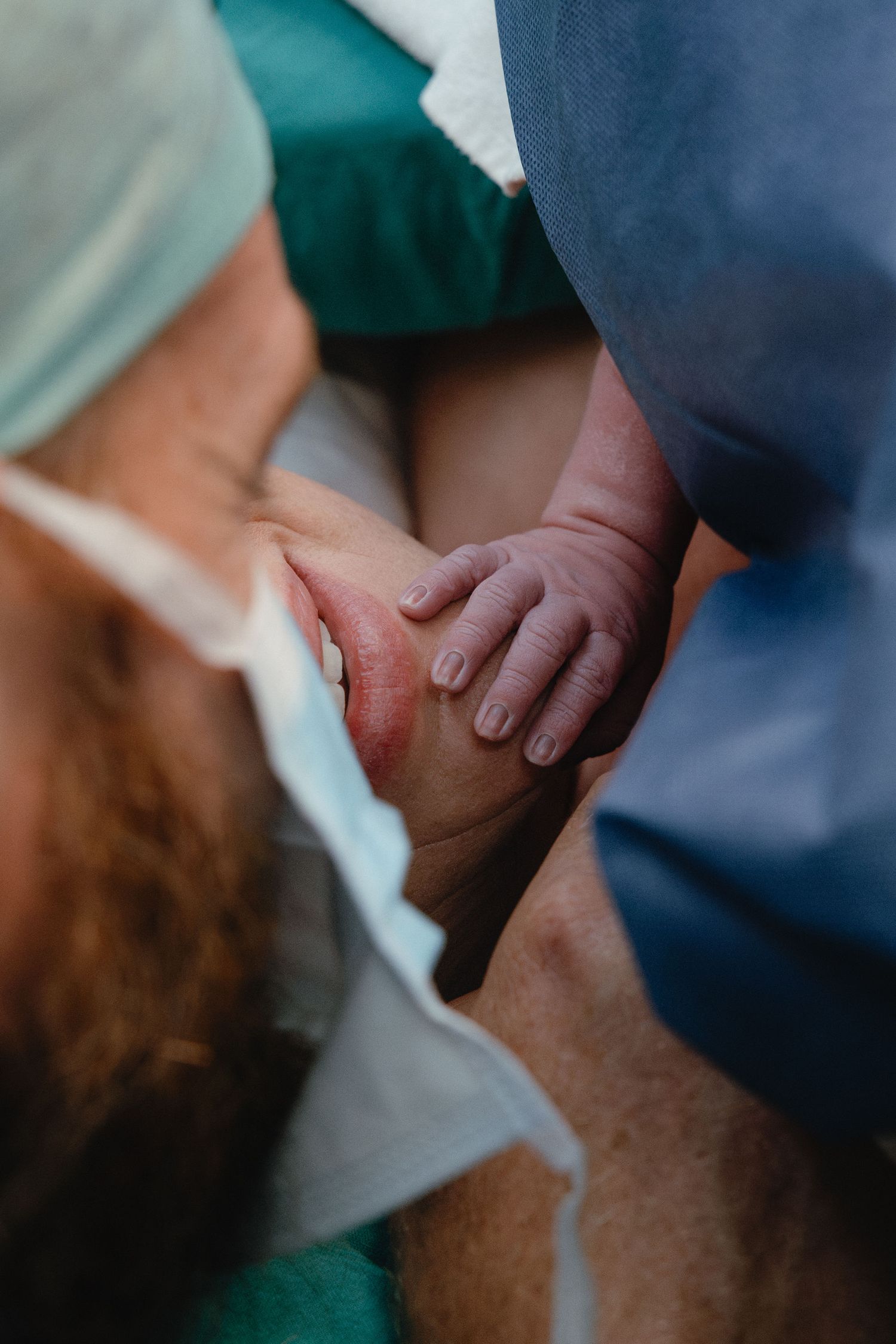 A newborn's tiny hand rests on a parent's leg as a bearded person in a surgical cap and mask leans close in a hospital delivery room.