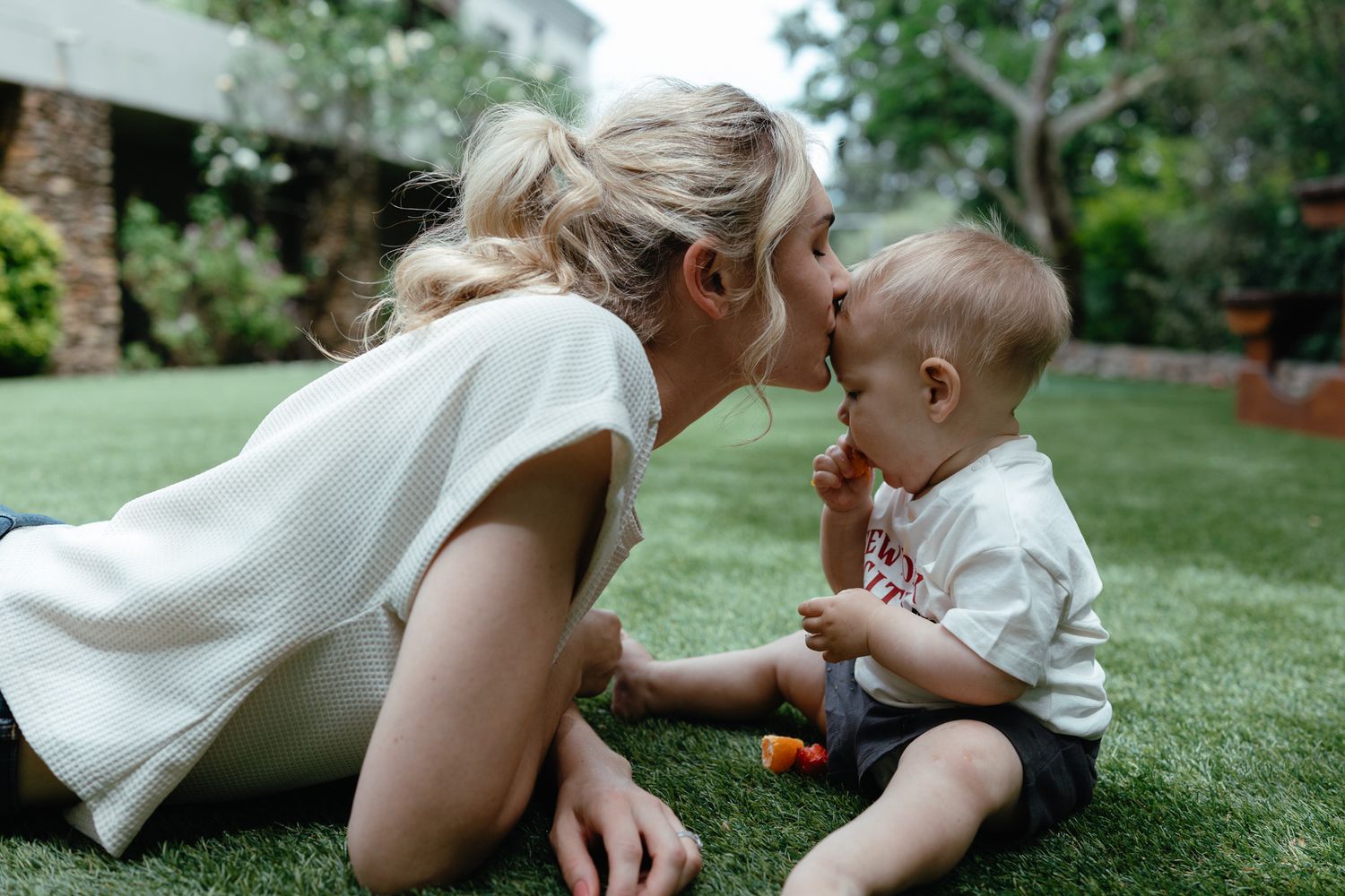 Mother kisses her smiling baby on the cheek while they sit together on a sunny backyard lawn.