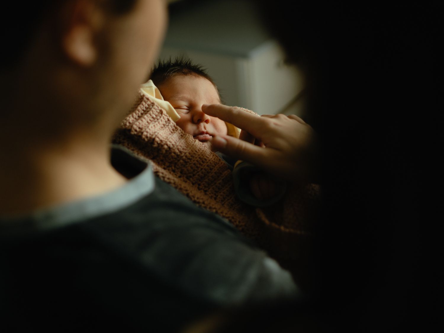 Parent gently touches a swaddled newborn baby's nose in a dimly lit room, framed by dark shadows.