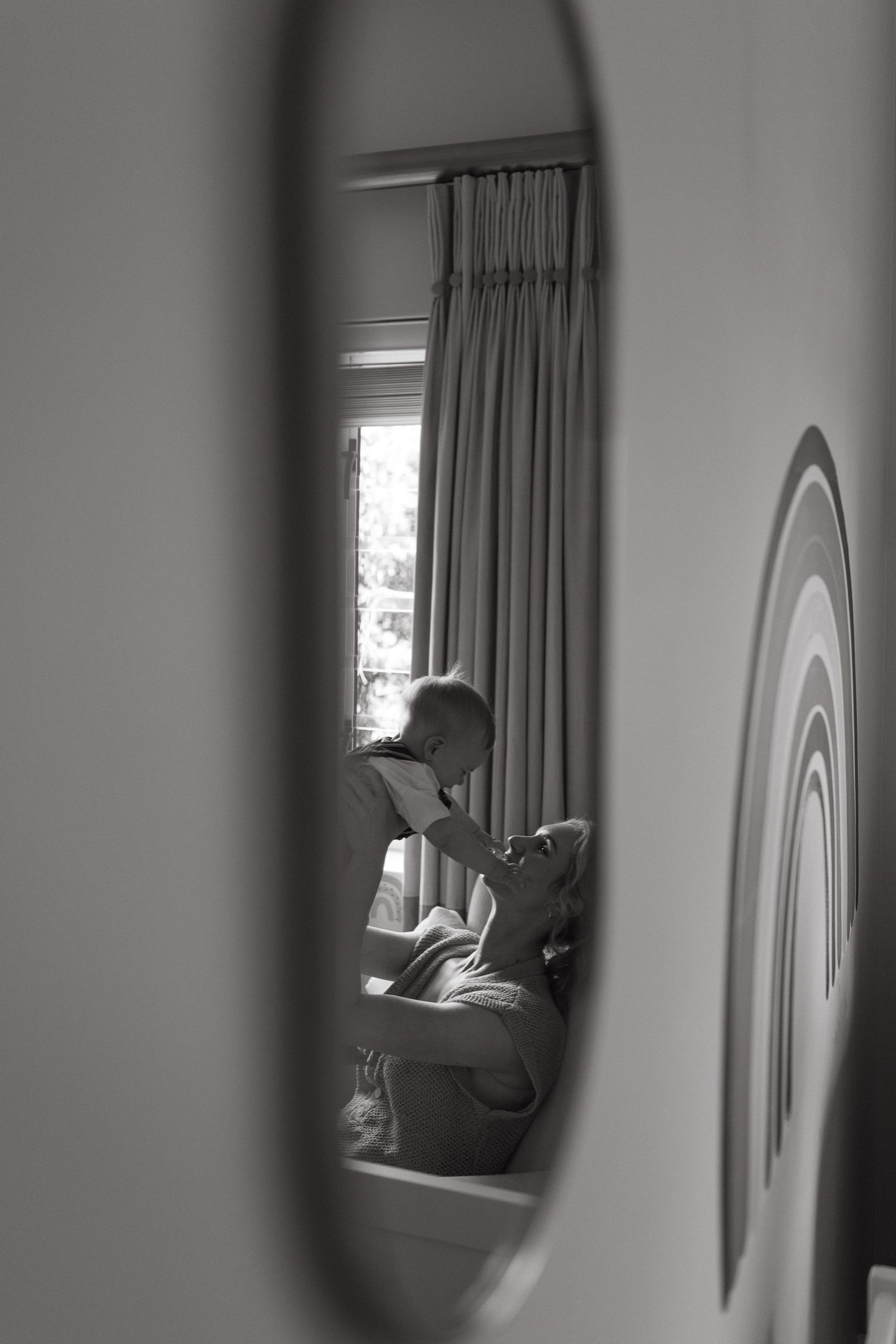 Black and white mirror reflection of a mother lying back as she lifts and plays with her baby by a window with curtains.
