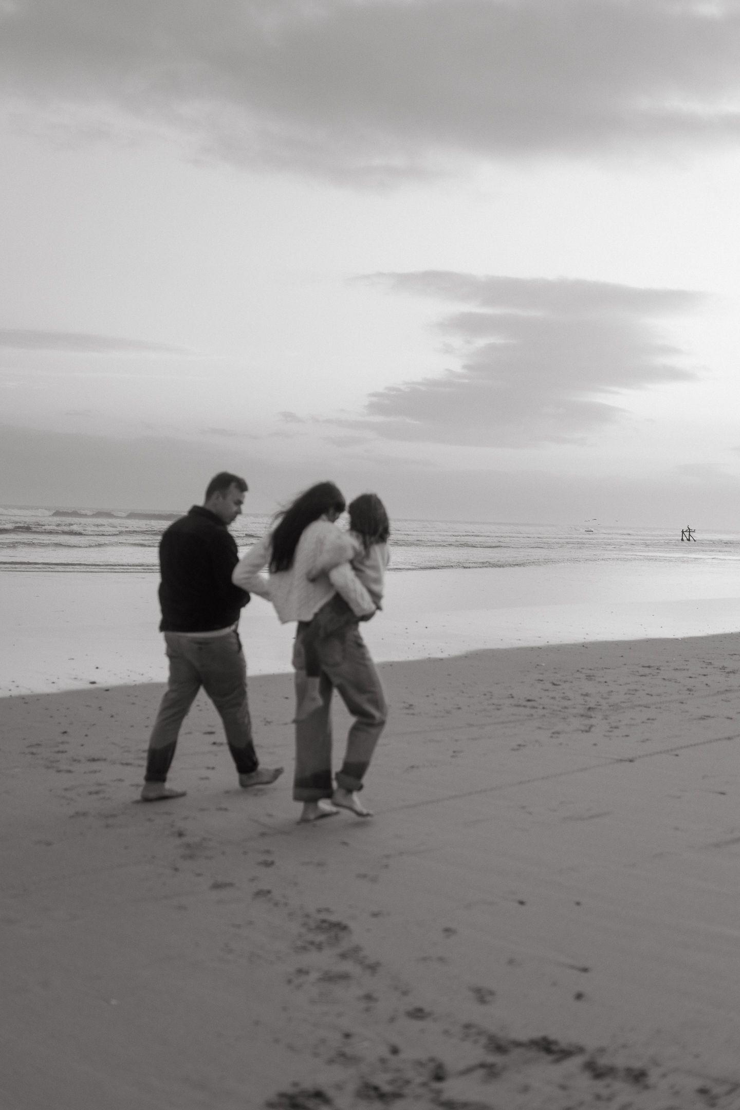 Black and white photo of a family of three walking along a quiet beach at dusk, with a parent carrying a child toward the waves.