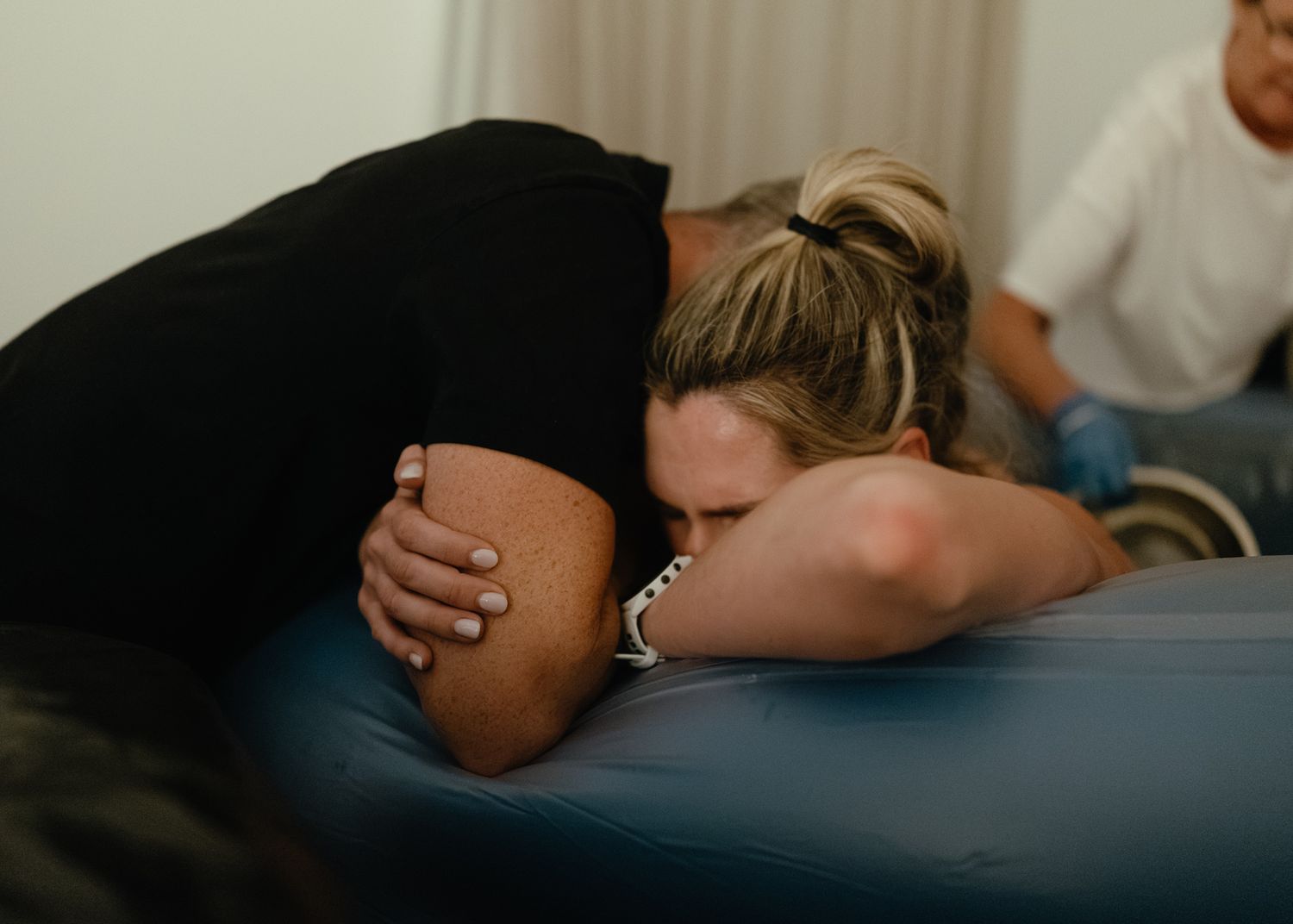 Woman in labor rests her head on her partner's arm while leaning over the edge of a birth pool as a midwife looks on.