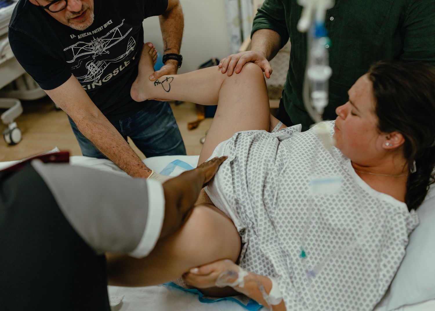 A laboring woman in a hospital gown lies on her side with an IV in her hand while her partner and a nurse support her raised leg.