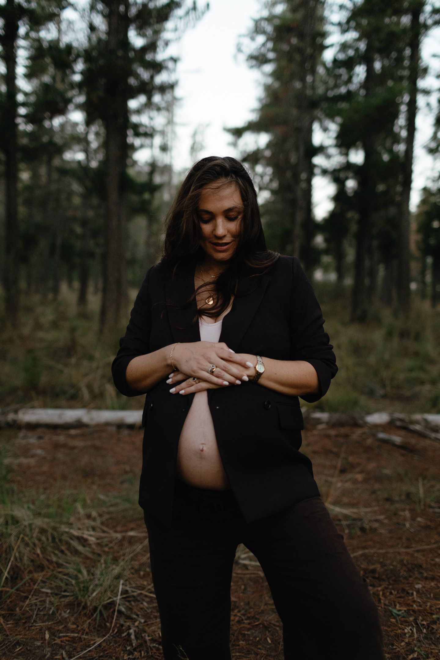 Pregnant woman in a black blazer gently holds her bare belly while standing among tall pine trees.