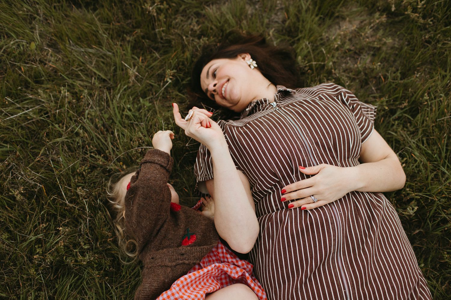 A woman in a striped dress lies on green grass beside a little girl, holding hands in a peaceful moment viewed from above.